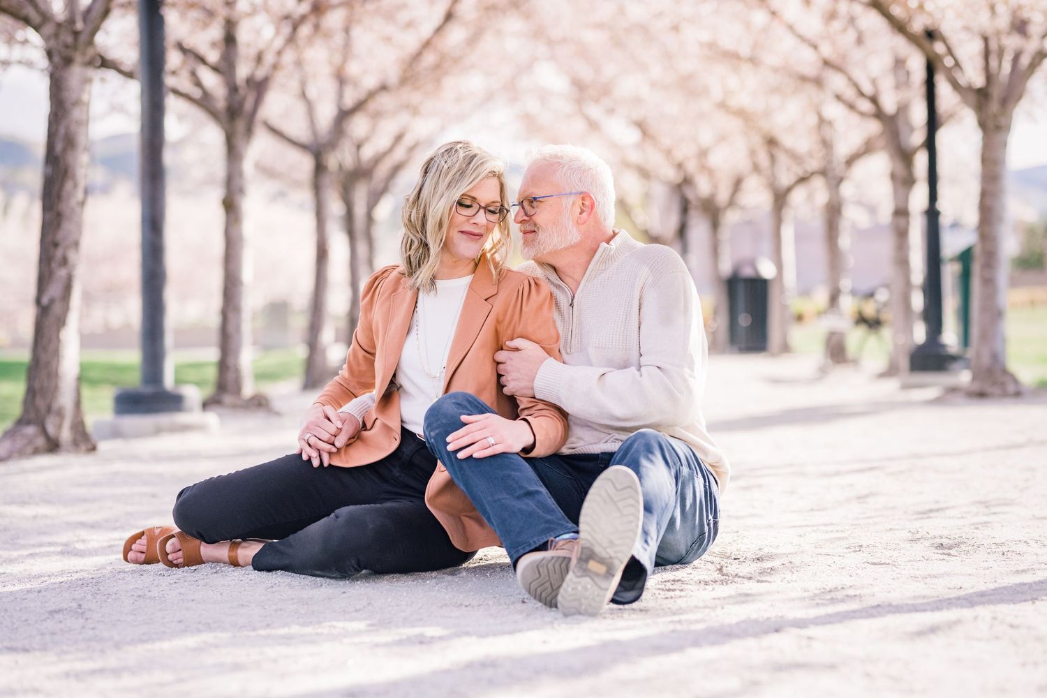A couple sits together on the ground surrounded by pink cherry blossom trees.
