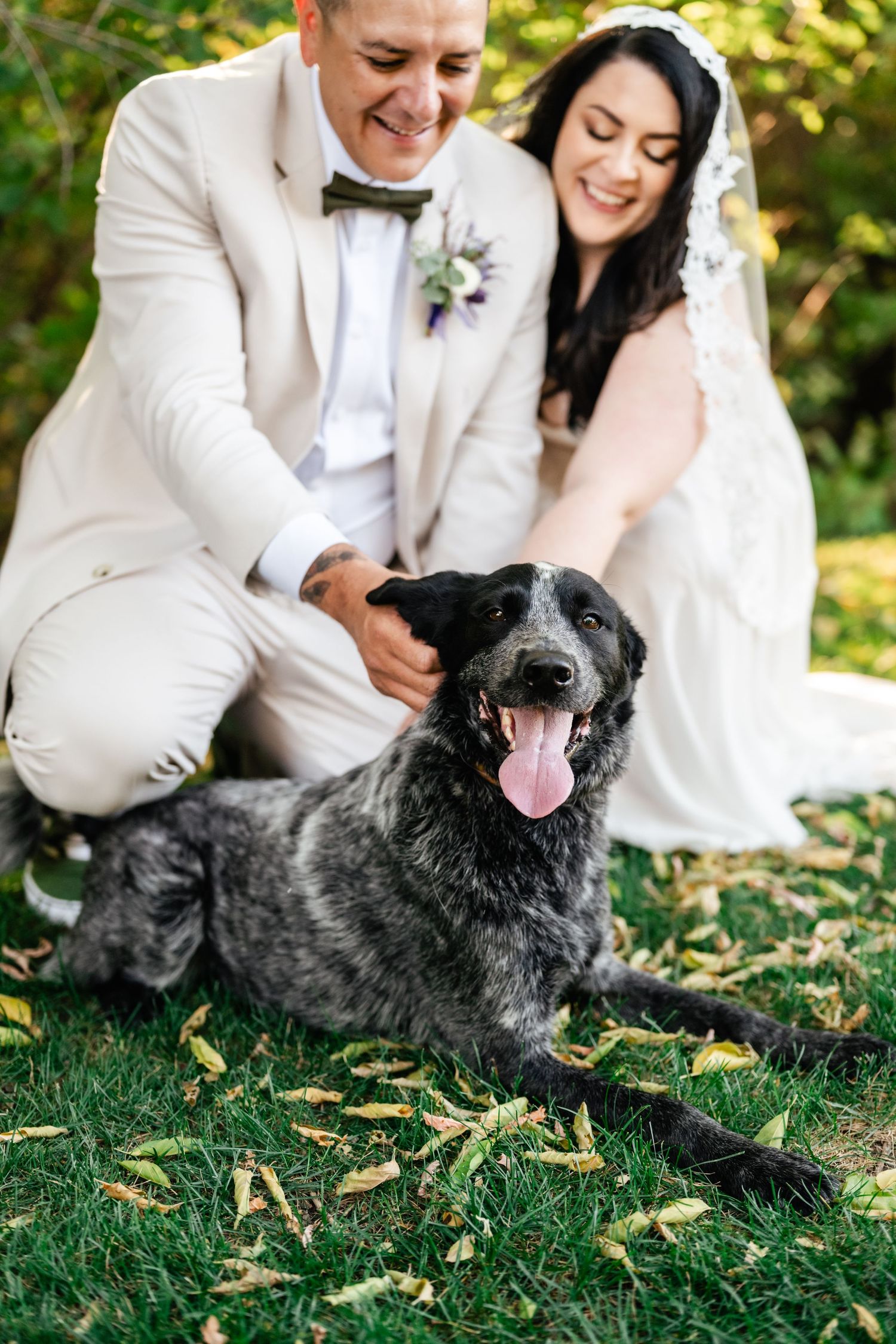 A happy couple in wedding attire sits on grass with their smiling black dog during an outdoor ceremony.