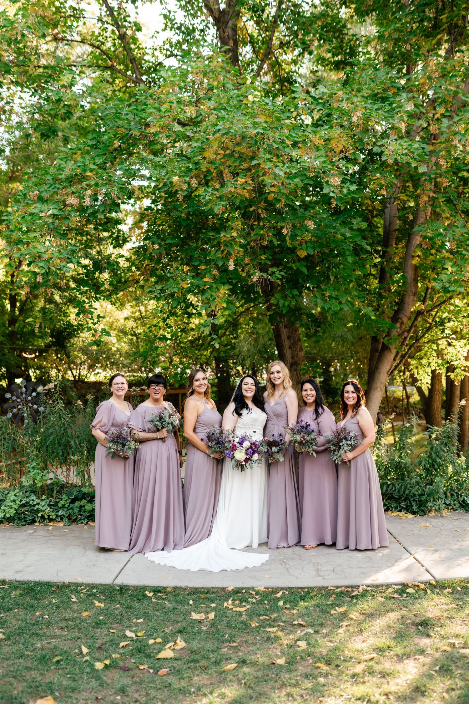 A bridal party in lavender dresses stands together on a garden path surrounded by lush green trees and foliage.
