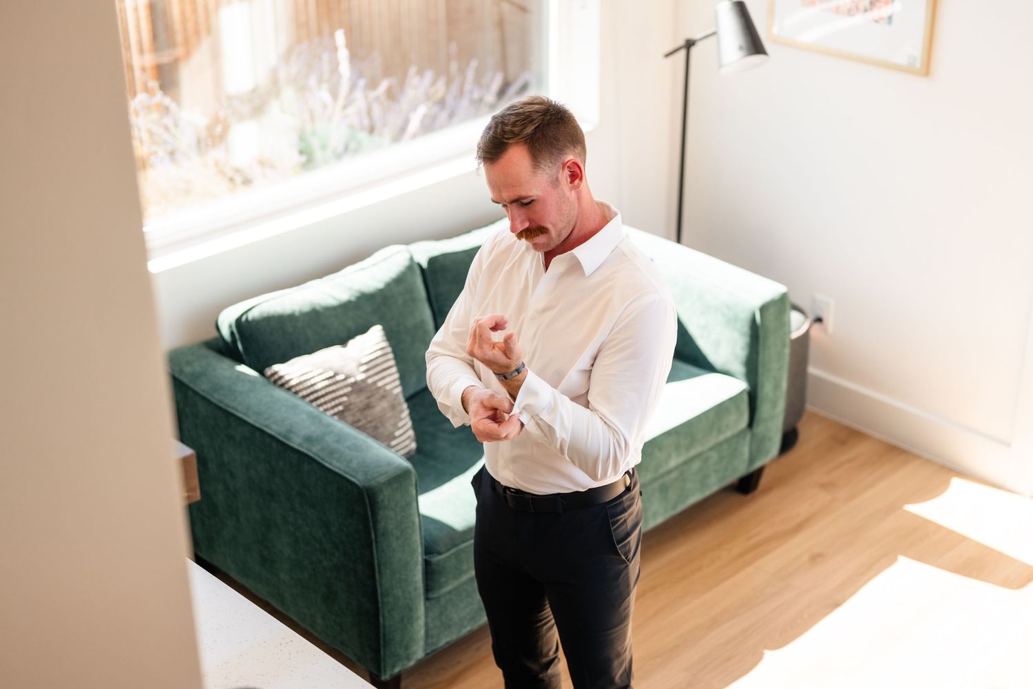 Person in white dress shirt adjusts clothing while standing near emerald velvet couch in bright room.