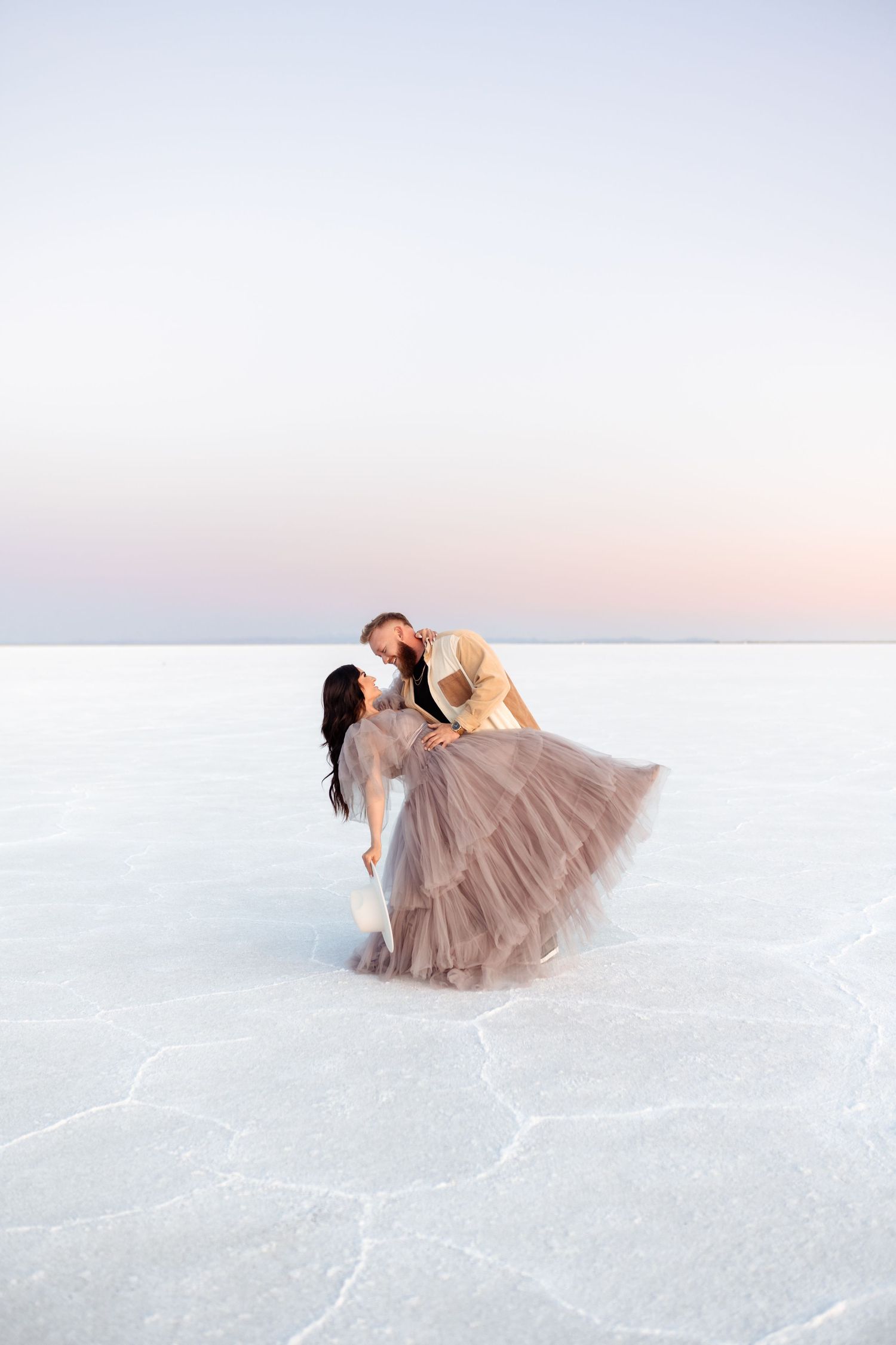 A couple in formal wear poses dramatically on a salt flat during sunset.