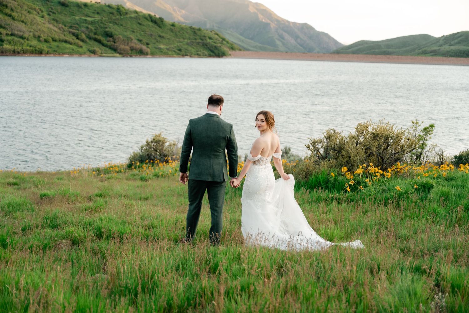 Bride in white gown and groom in green suit stand by mountain lake during sunset.