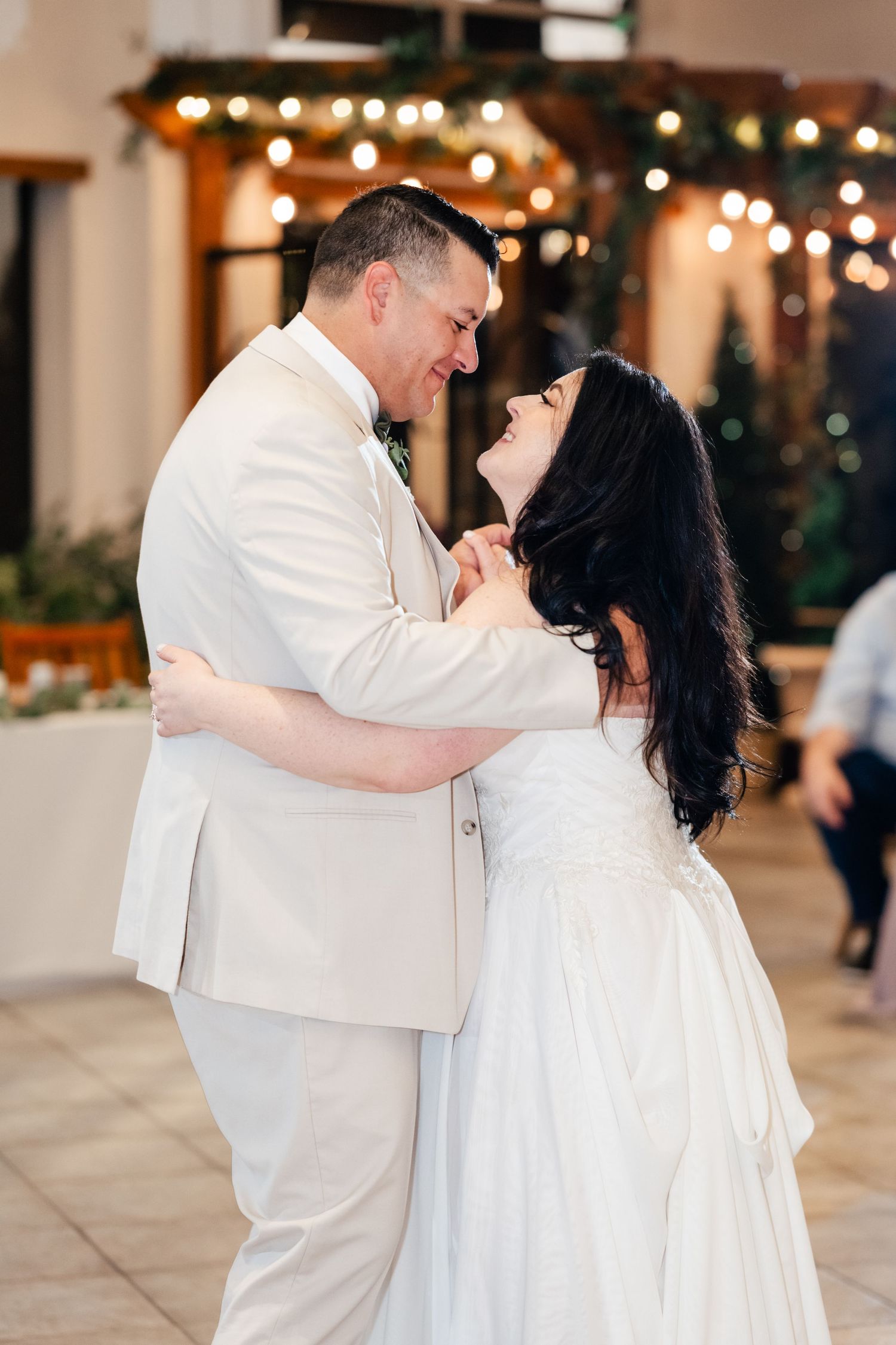 Couple dressed in white sharing an intimate dance moment at a wedding reception with twinkling lights in background.