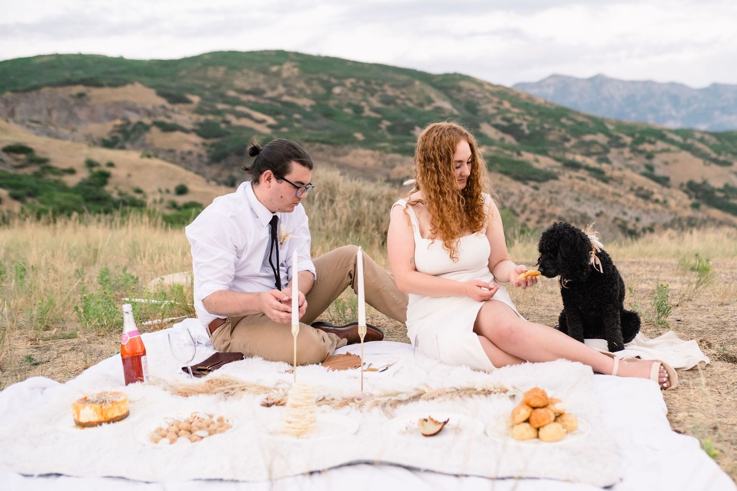 A group enjoying a romantic sunset picnic with wine and snacks on a blanket overlooking rolling hills and mountains. Utah mountain elopement