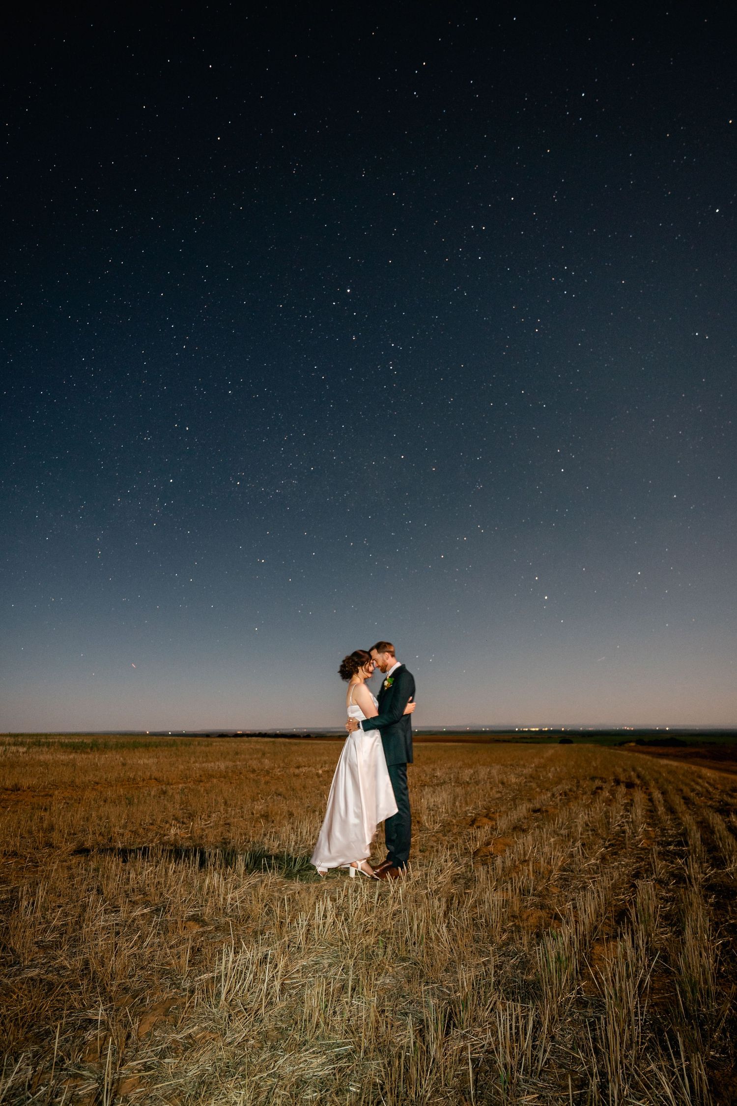 Wedding couple embracing under starlit sky in vast wheat field at dusk.