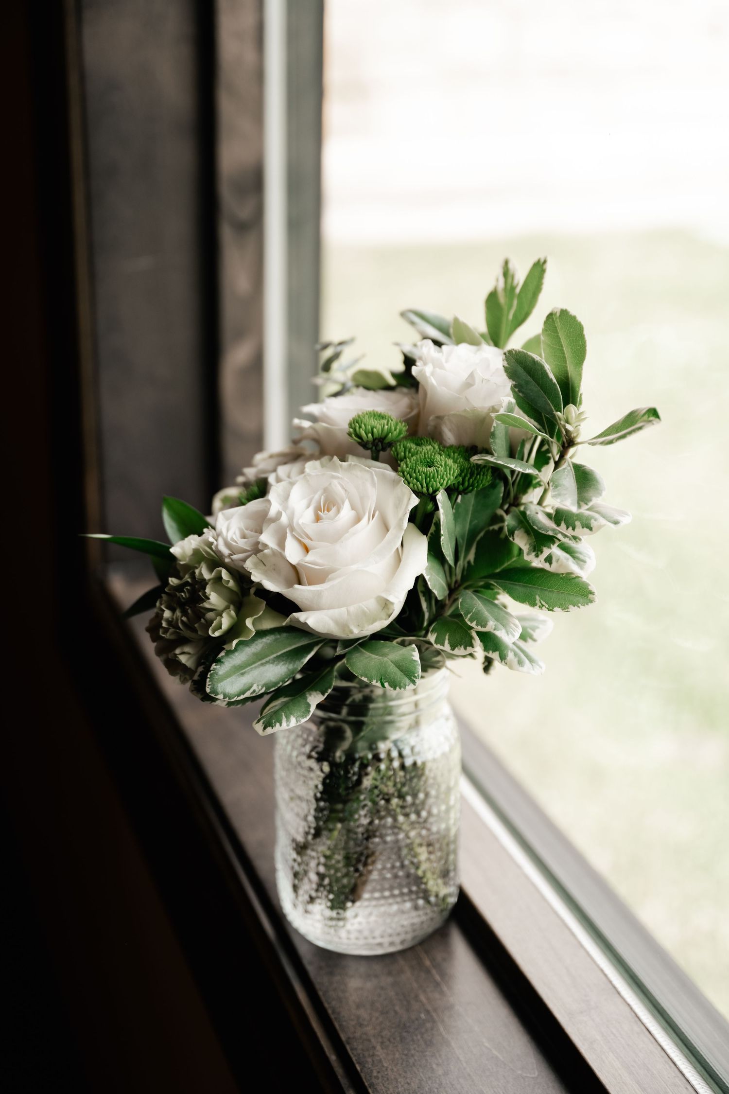 White roses and greenery arranged in a glass vase sitting on a windowsill in natural light.