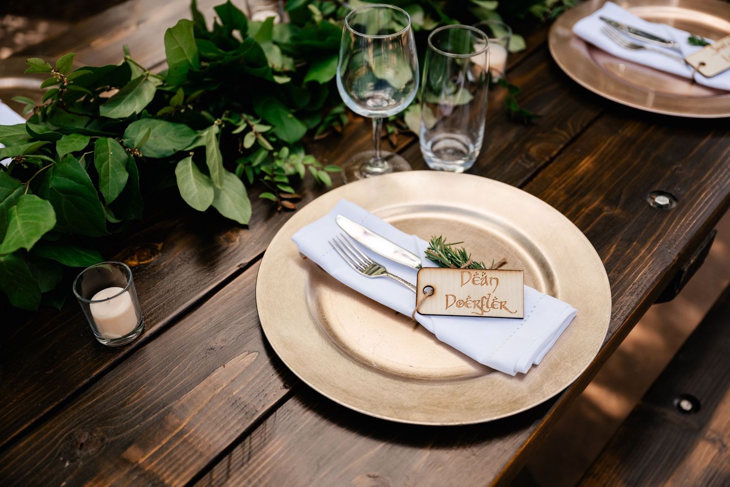 Rustic wooden table set for dining with white plates, wine glasses, and green foliage decor.