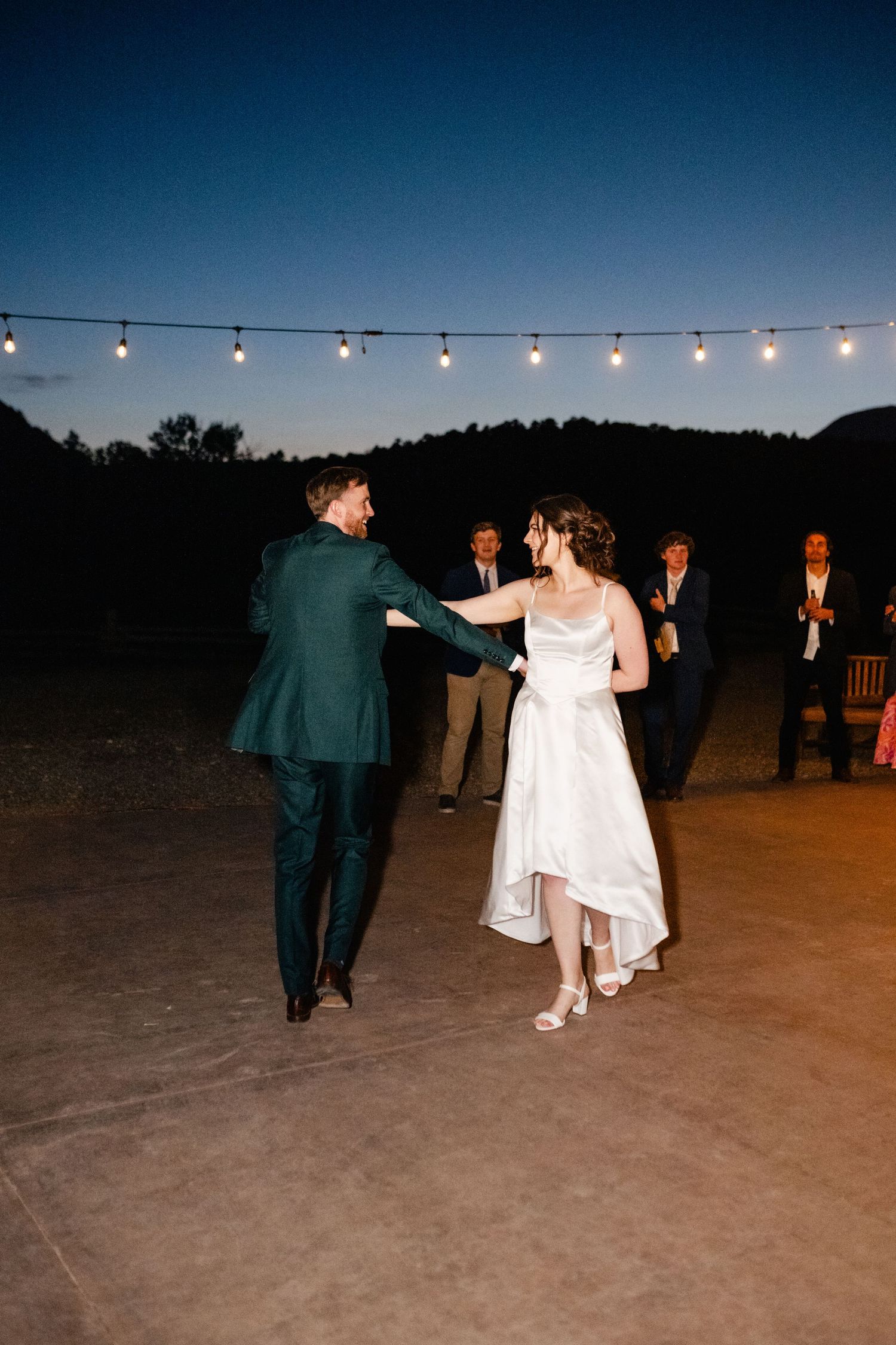 Couple dances under string lights at outdoor evening wedding reception.