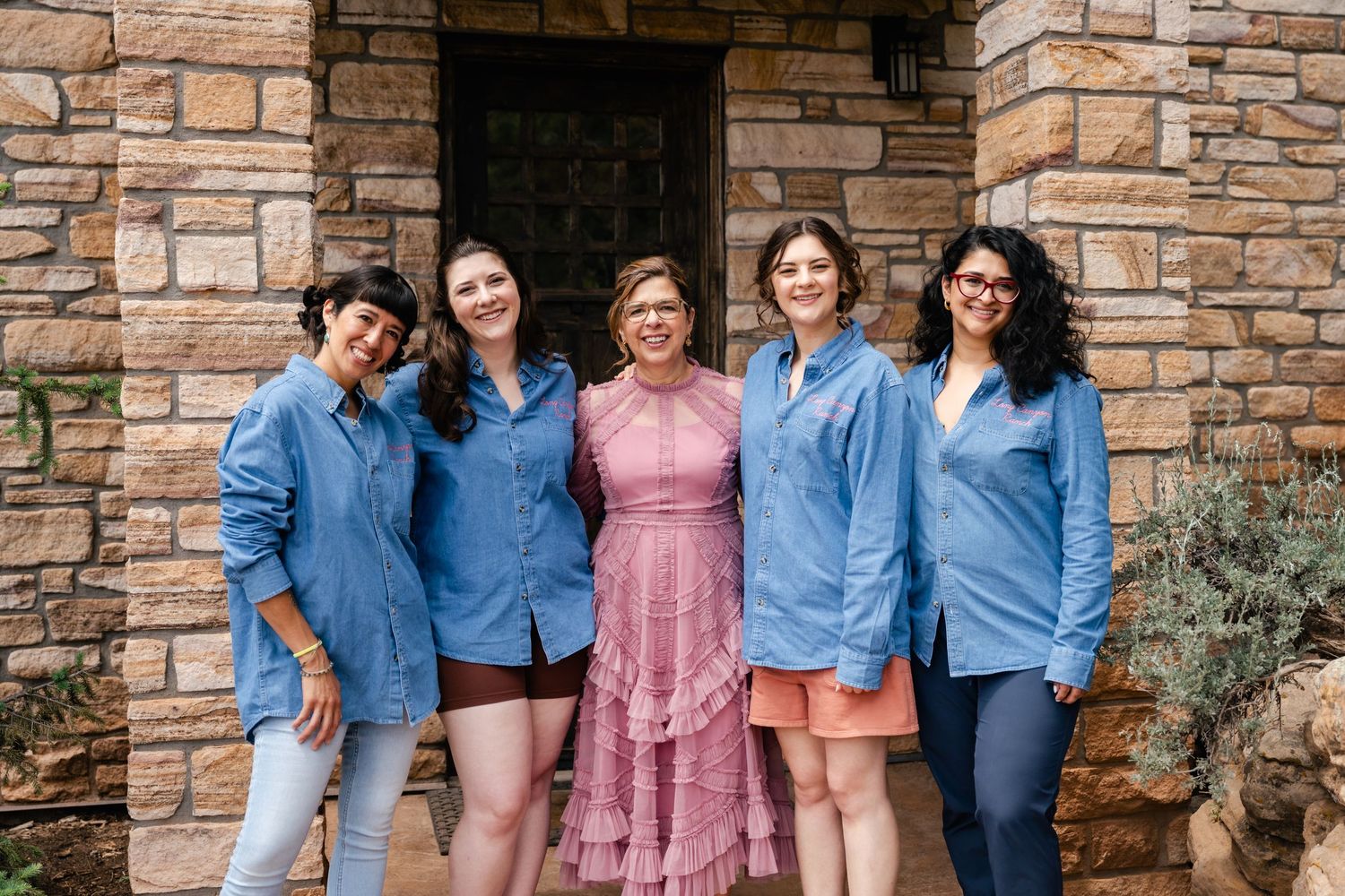 A group in coordinated blue and pink outfits pose against a stone wall exterior.