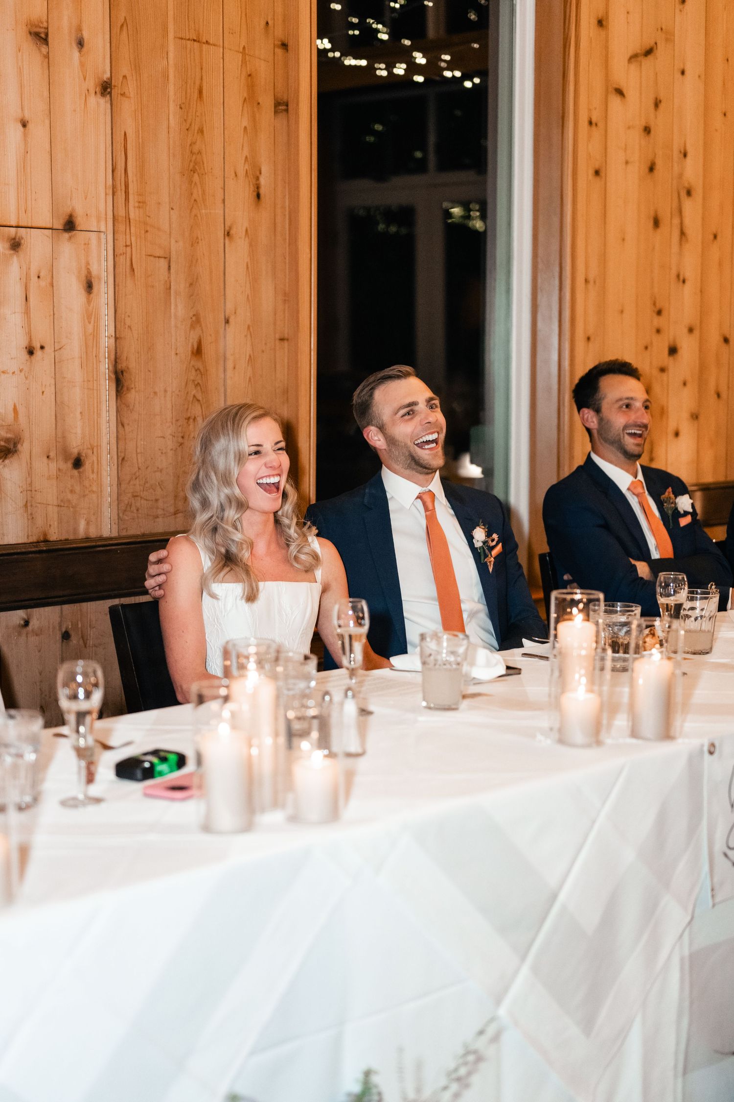 Wedding reception guests seated at long tables with orange accents and candlelight in a rustic wooden venue.