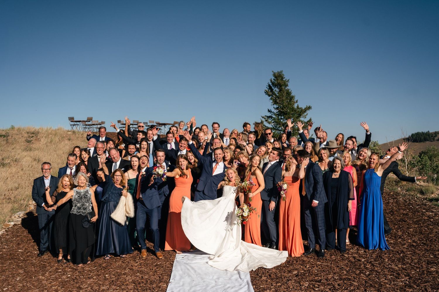 Wedding celebration group photo with guests arranged on hillside against mountain scenery.