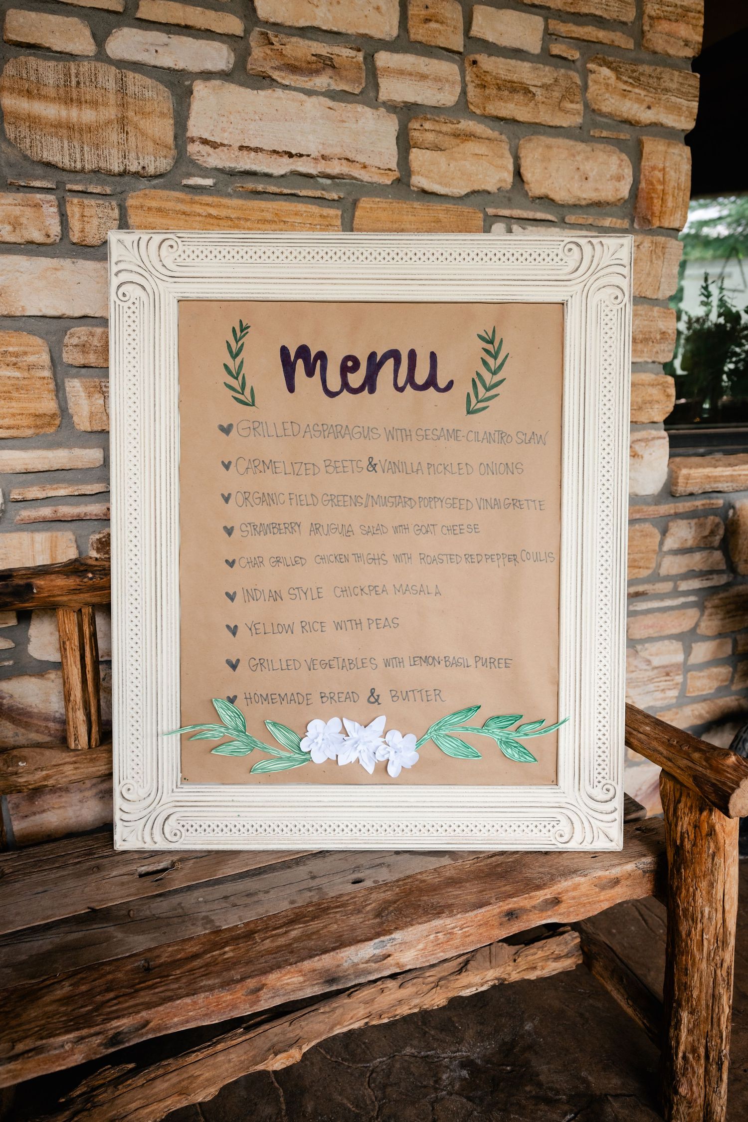 Rustic wedding menu displayed on kraft paper with floral decorations mounted on stone wall.
