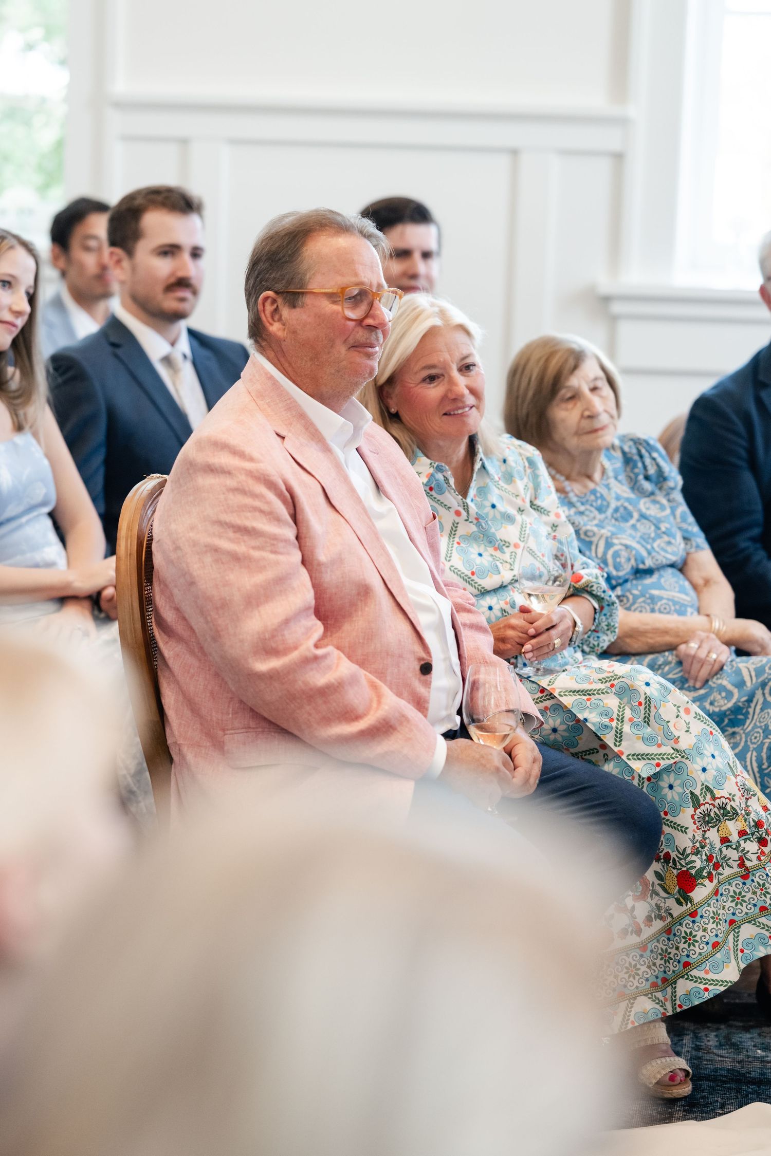 Wedding guests in floral and pink attire seated in rows during an indoor ceremony.
