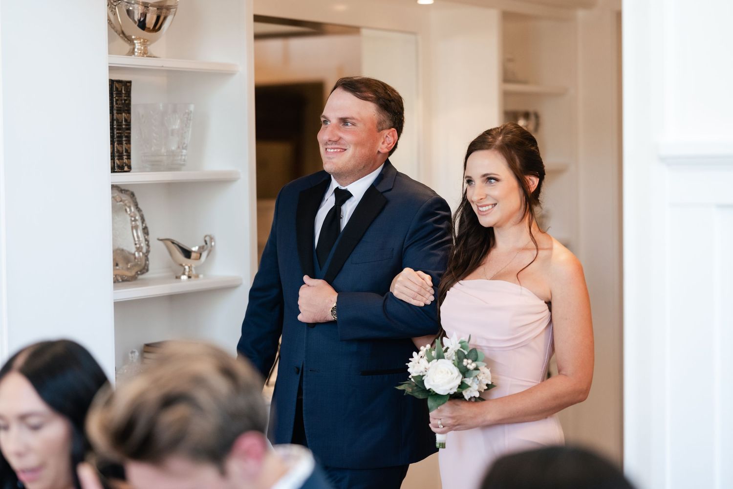 A couple in formal wedding attire walks down a hallway with white walls and built-in shelves.