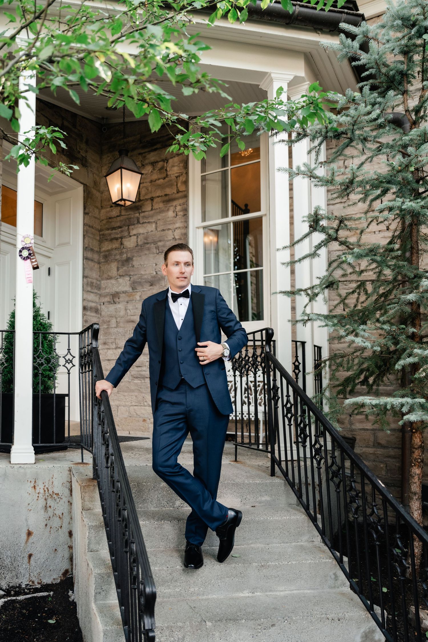 A groom in a navy tuxedo poses elegantly on stone steps with white columns and greenery in the background.