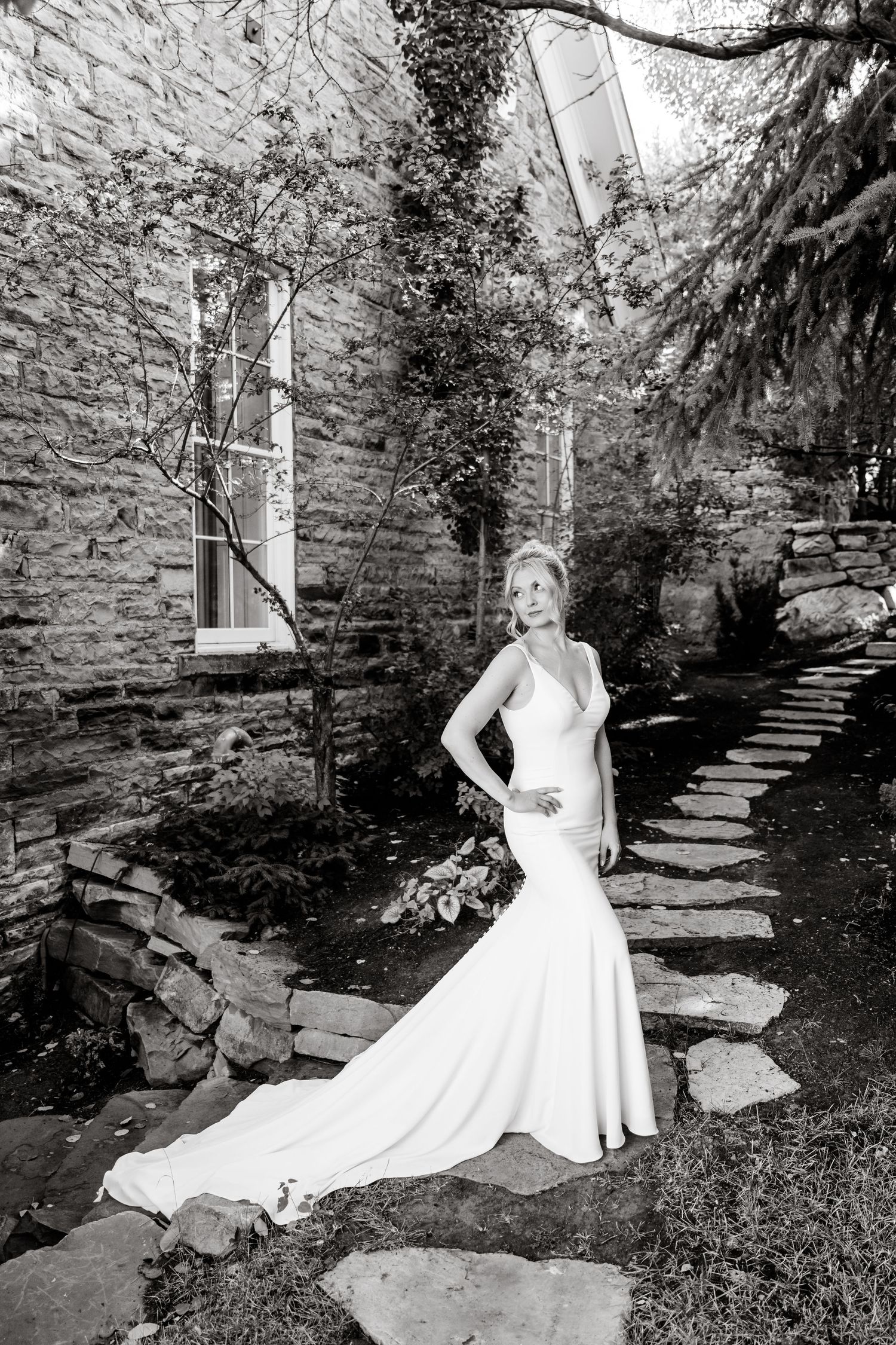 A stunning wedding dress with a mermaid silhouette and long train photographed against a rustic stone wall and garden path.