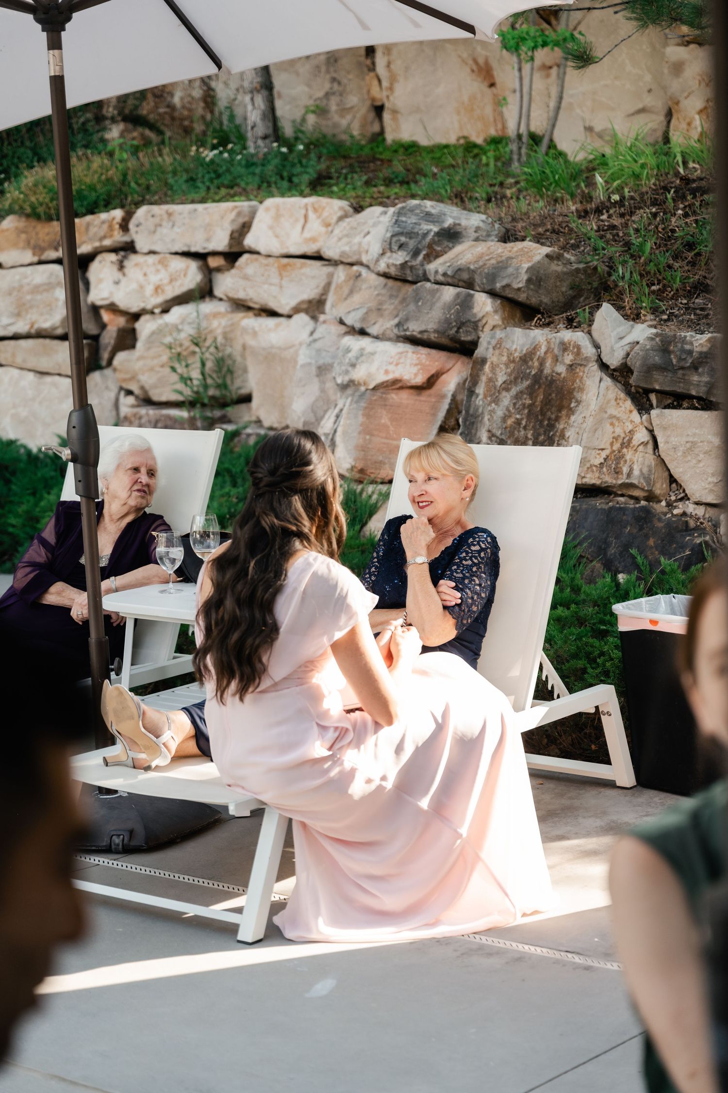Wedding guests relax in white lounge chairs against a stone wall backdrop during an outdoor reception.