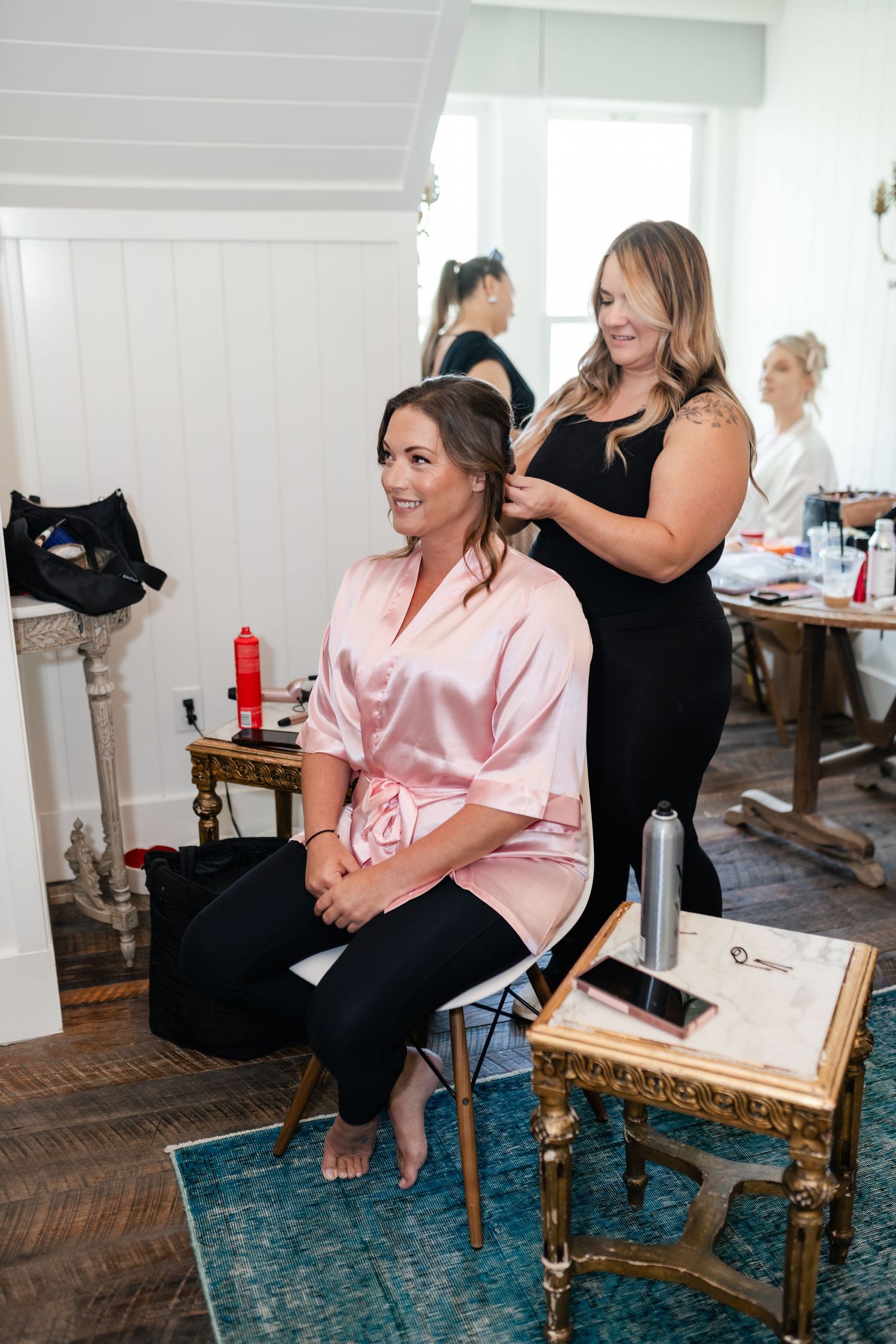 Stylists prepare someone wearing a pink silk robe in an elegant room with vintage furniture and blue carpet.