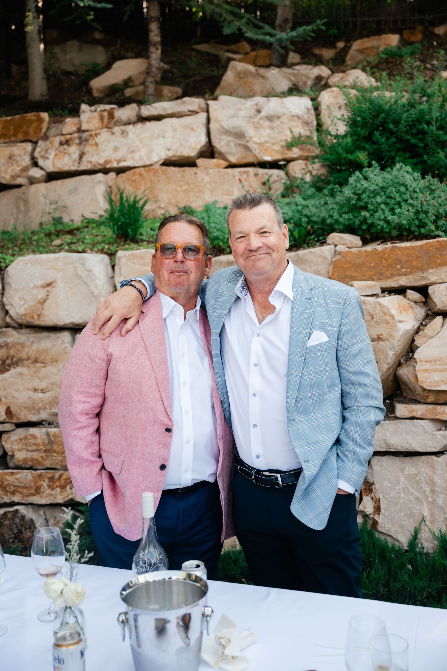 Wedding guests in formal attire gathered at an outdoor stone wall reception area.