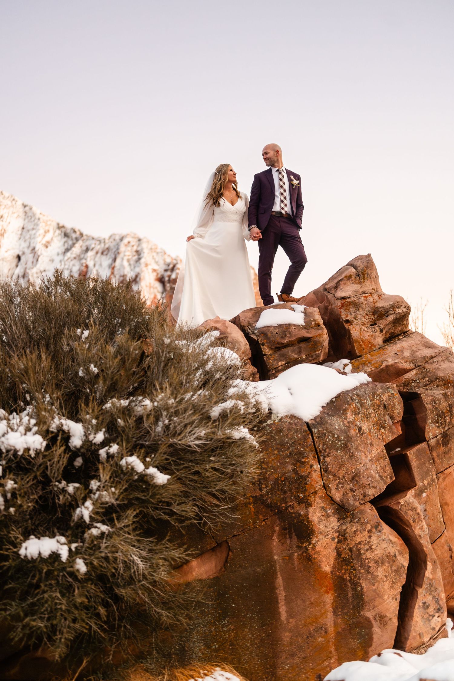 A couple poses dramatically on snow-dusted red rocks during sunset.