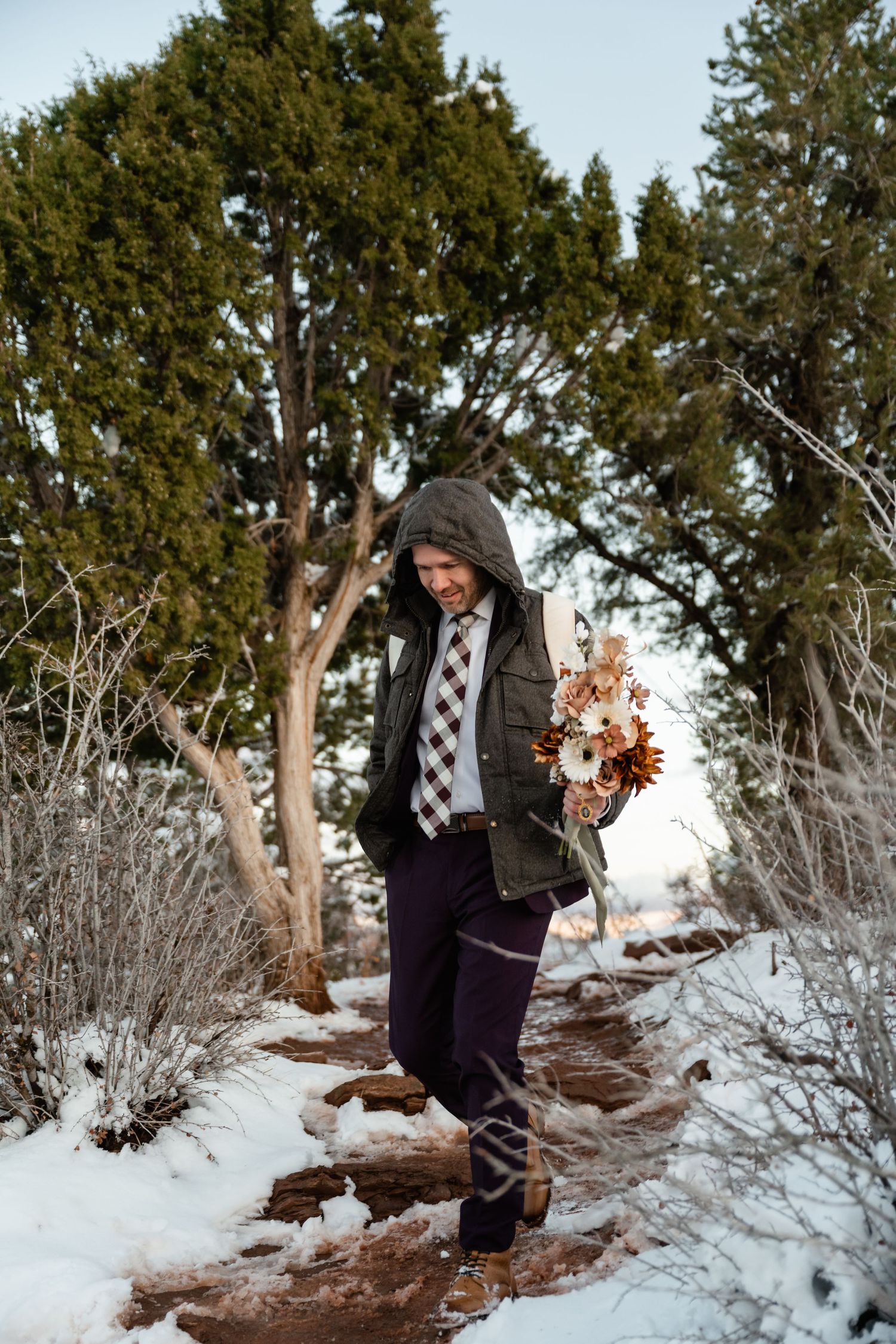 Two people walking along a snowy mountain trail surrounded by evergreen trees and winter vegetation.