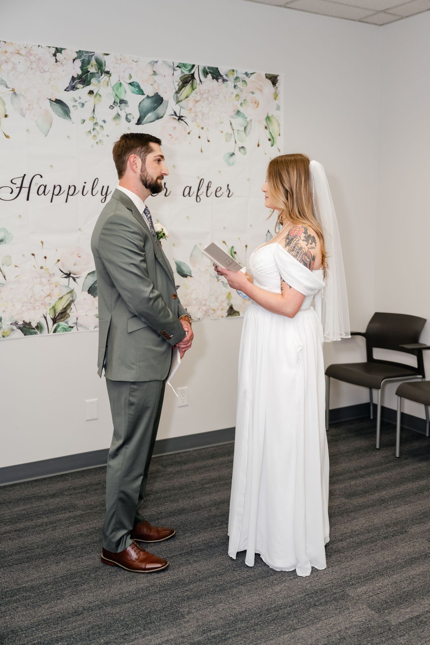 A couple exchanges vows in front of a floral backdrop with 'Happily Ever After' text during an intimate wedding ceremony.