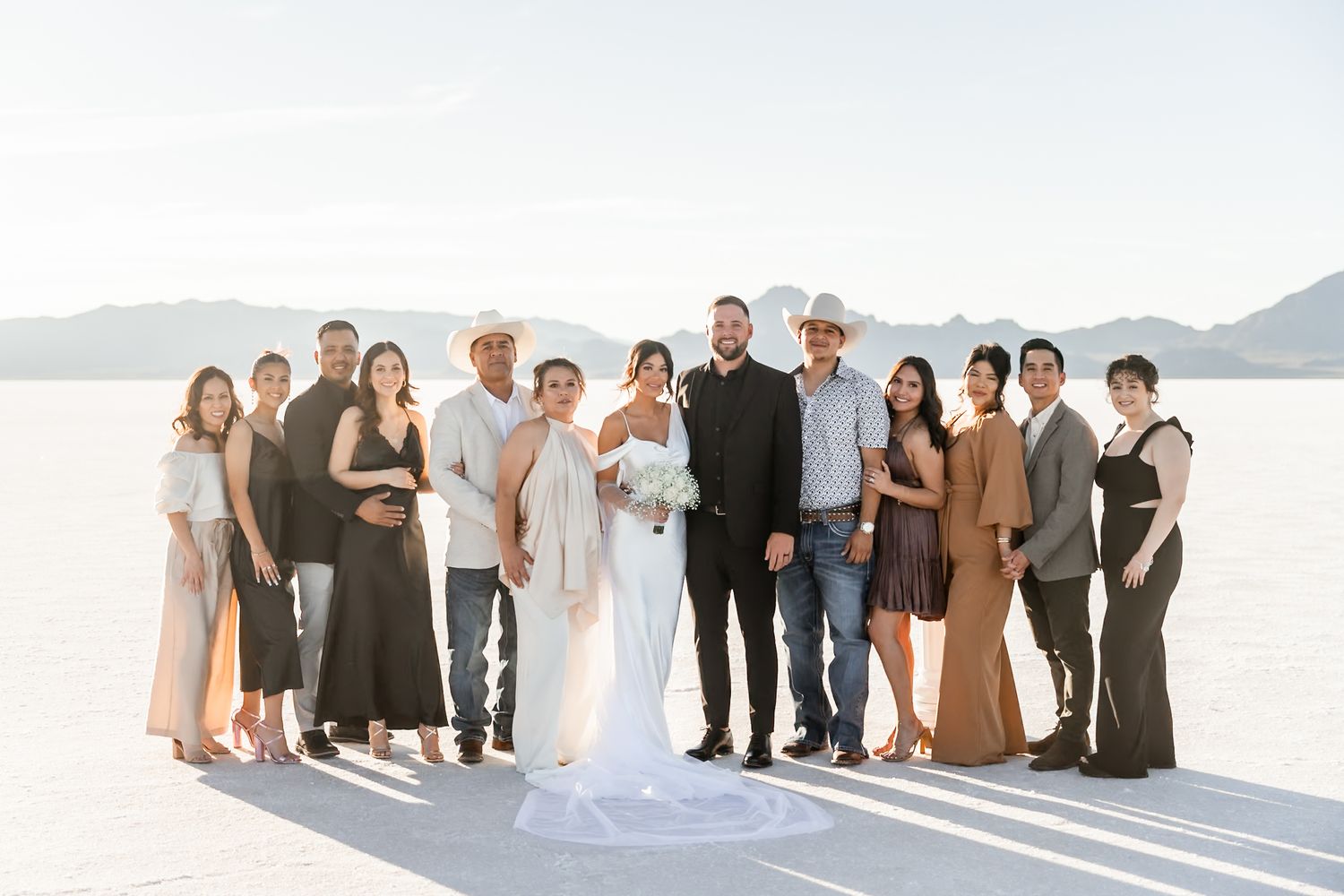 A wedding party in neutral-toned attire poses together on a bright white salt flat with mountains in the background.