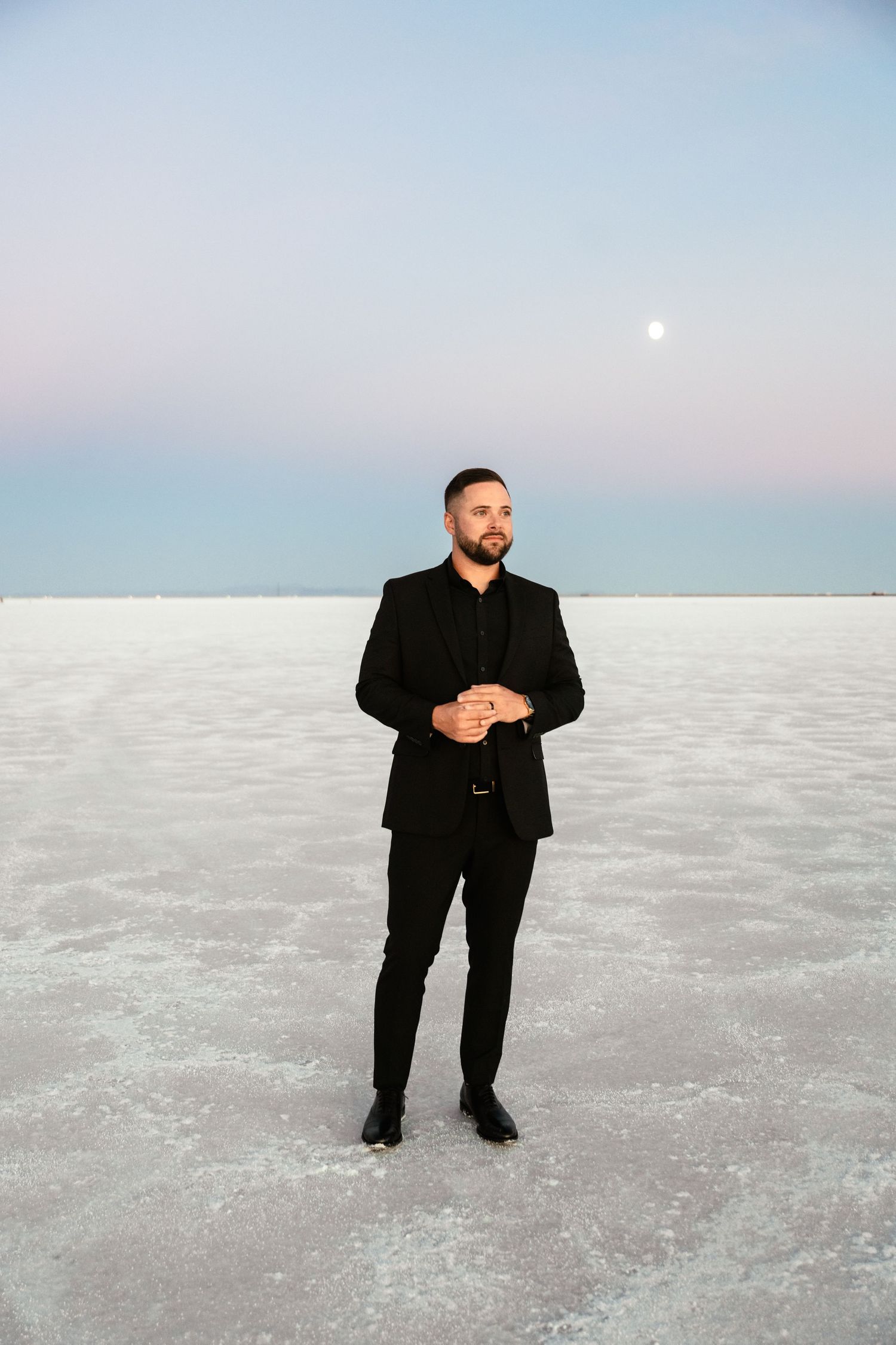 A figure in formal black attire poses on white salt flats under a pastel sky with visible moon.