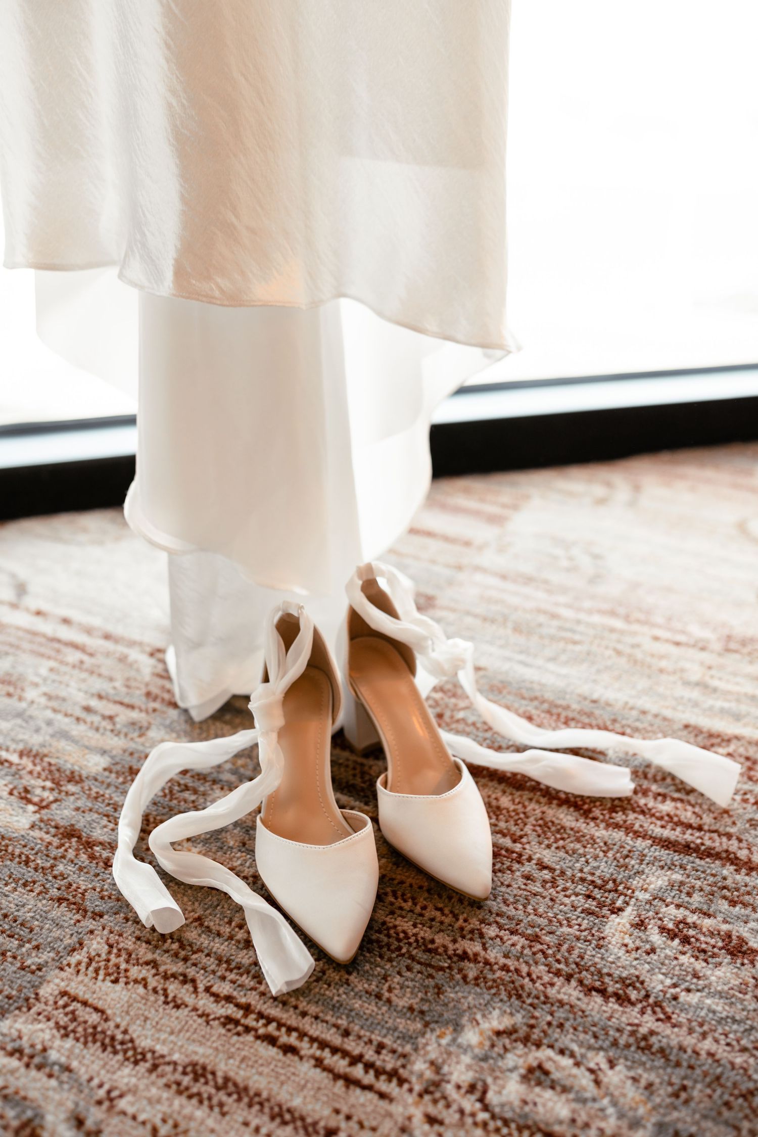 Elegant white bridal shoes with bows displayed on a patterned carpet.