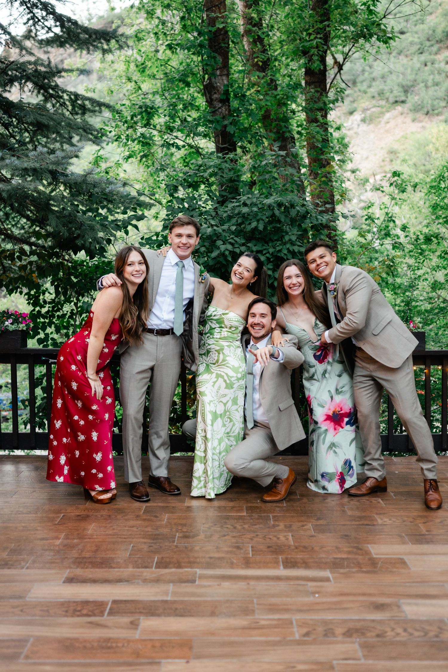 Wedding party poses playfully on wooden deck surrounded by lush green forest during outdoor celebration. Millcreek Inn Wedding Photographer.