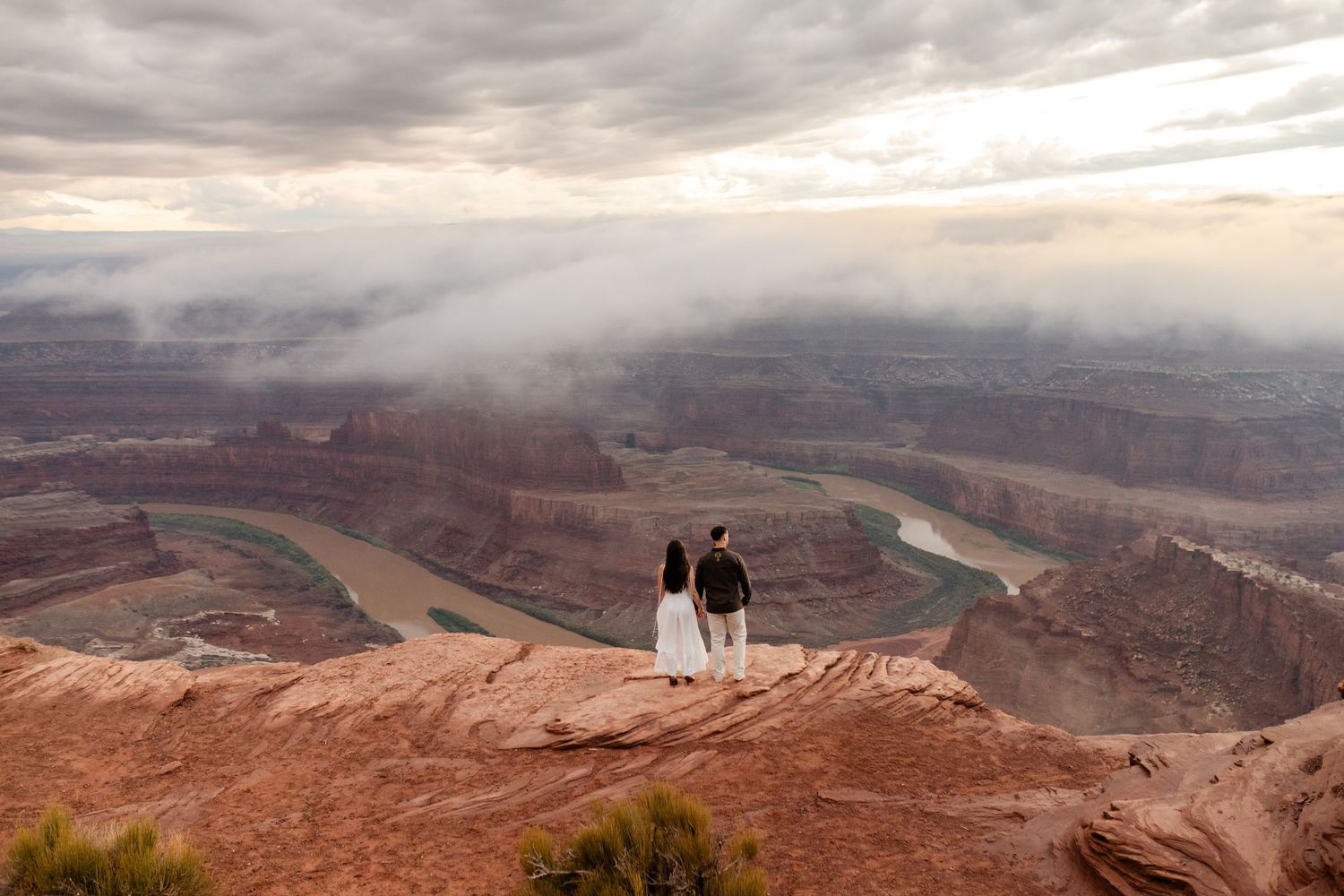 Two silhouettes stand on a cliff edge overlooking a vast canyon landscape during sunset.