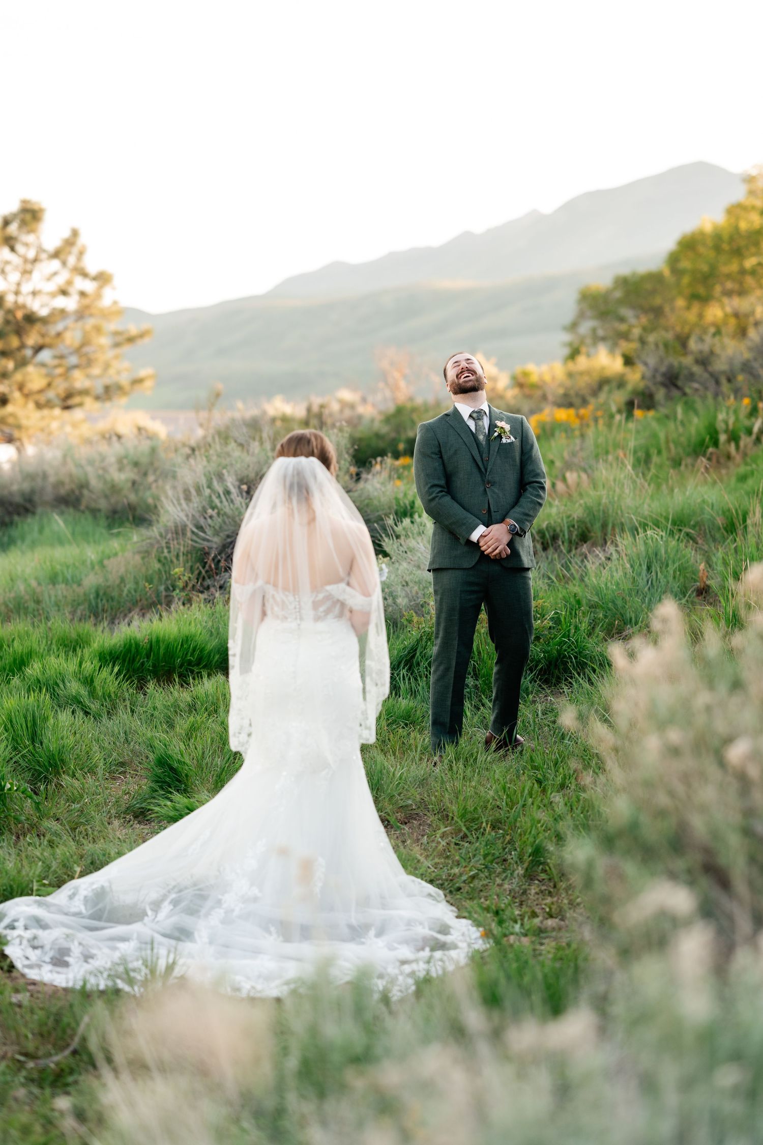A wedding couple poses in a scenic mountain meadow with a flowing white veil and dark suit at sunset.