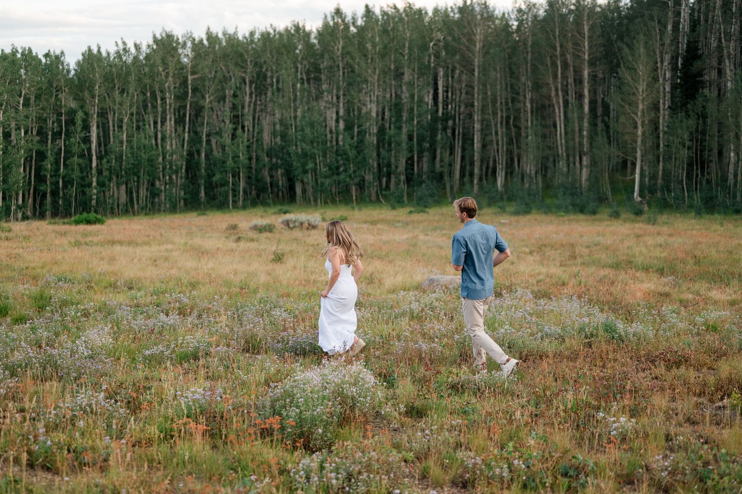 Couple running through a wildflower meadow with a lush green forest backdrop during their mountain engagement near Park City.
