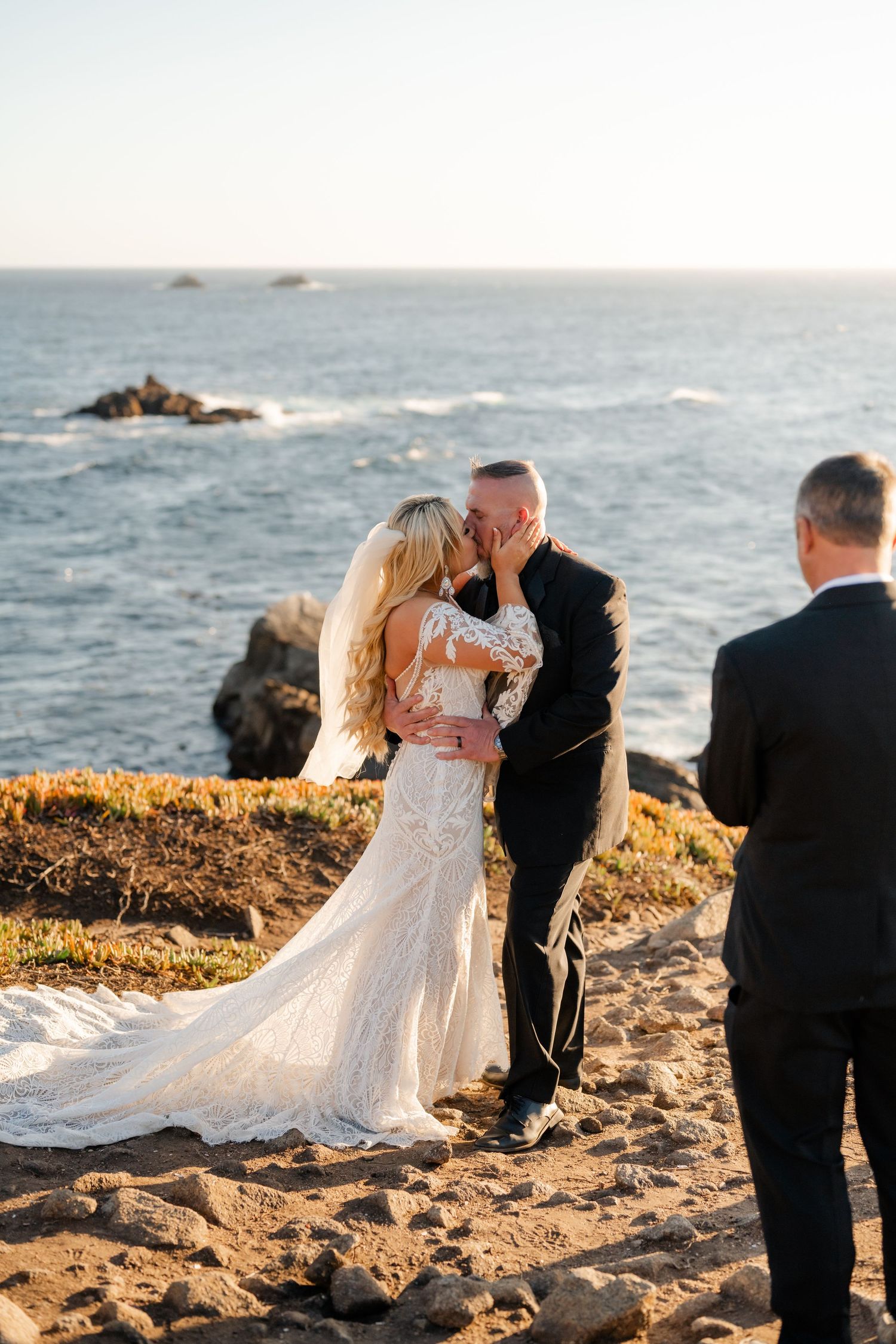 A romantic coastal wedding ceremony takes place at sunset with waves crashing against rocky cliffs in the background.