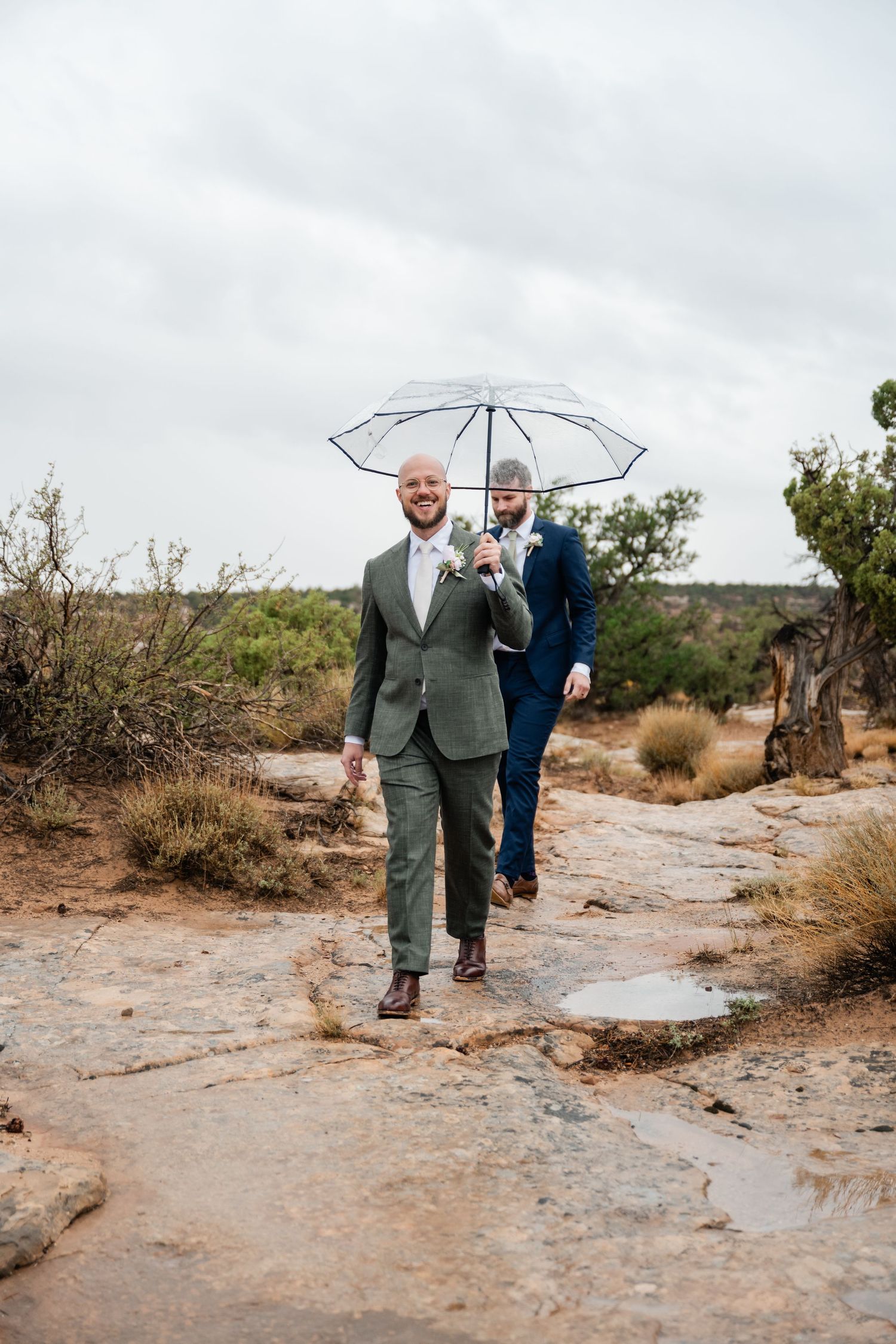 Two people walking on a rocky trail holding a clear umbrella against an overcast sky.