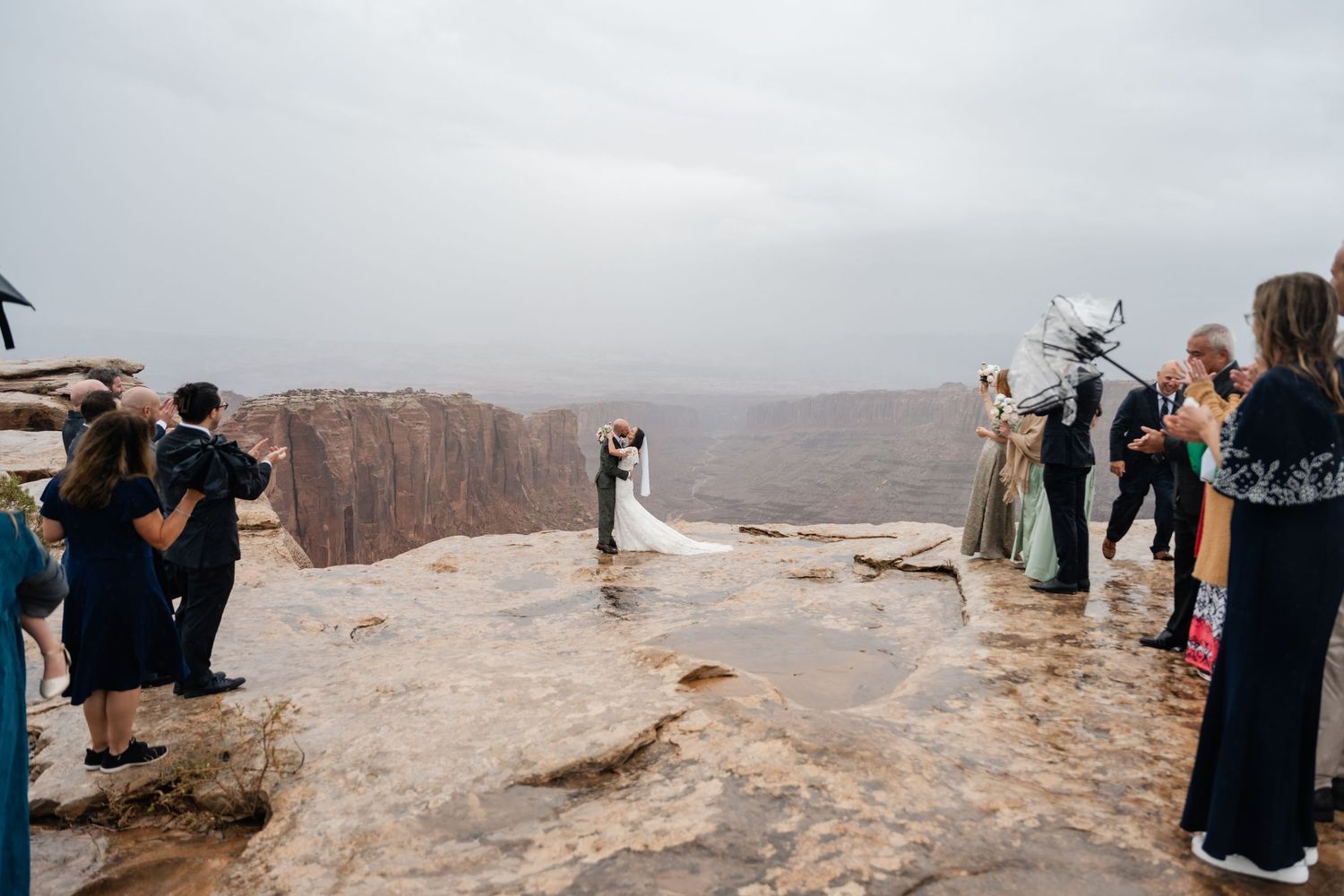 Panoramic view of wedding guests gathered on rocky cliff edge under cloudy skies.