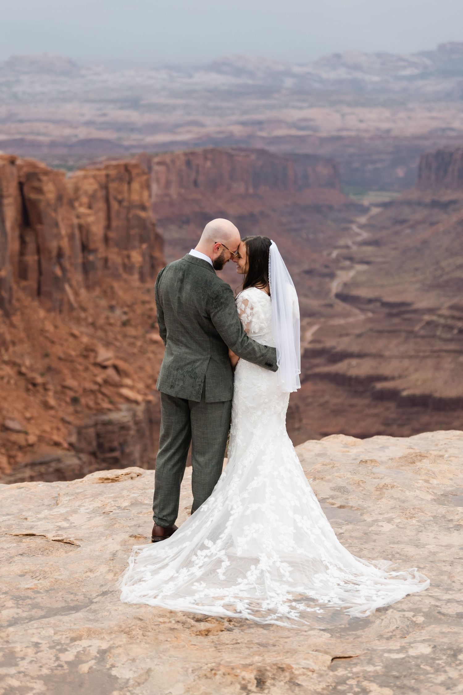 A wedding couple embraces on the edge of a scenic desert canyon with flowing white dress and dramatic landscape.
