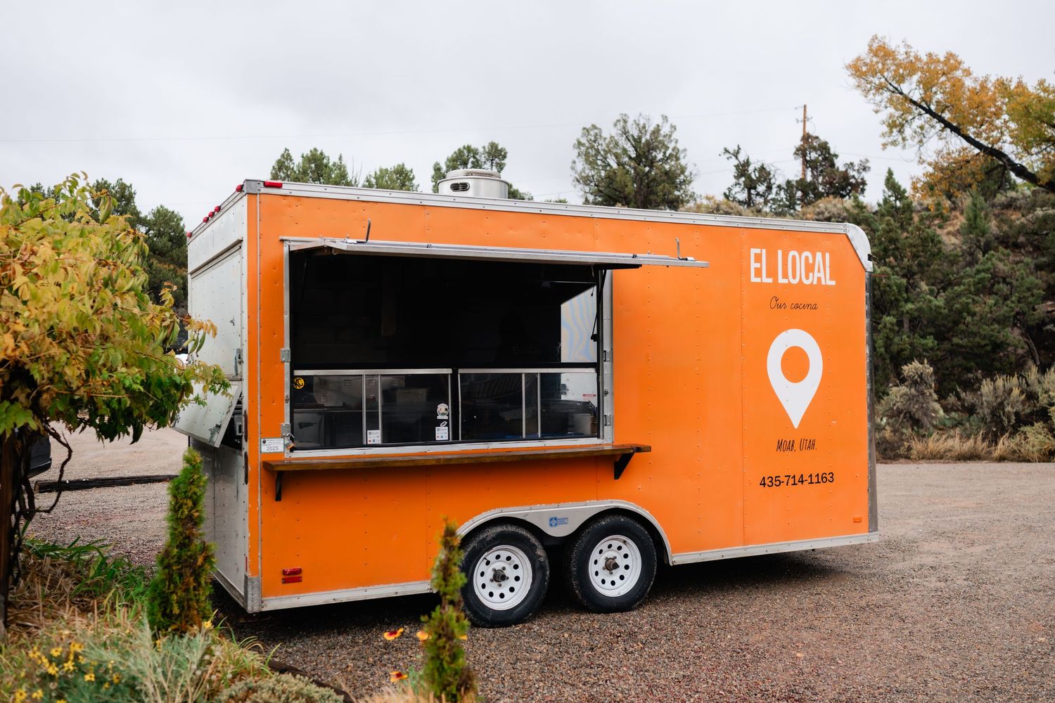 A bright orange food trailer with a service window parked in an outdoor setting with trees in the background.