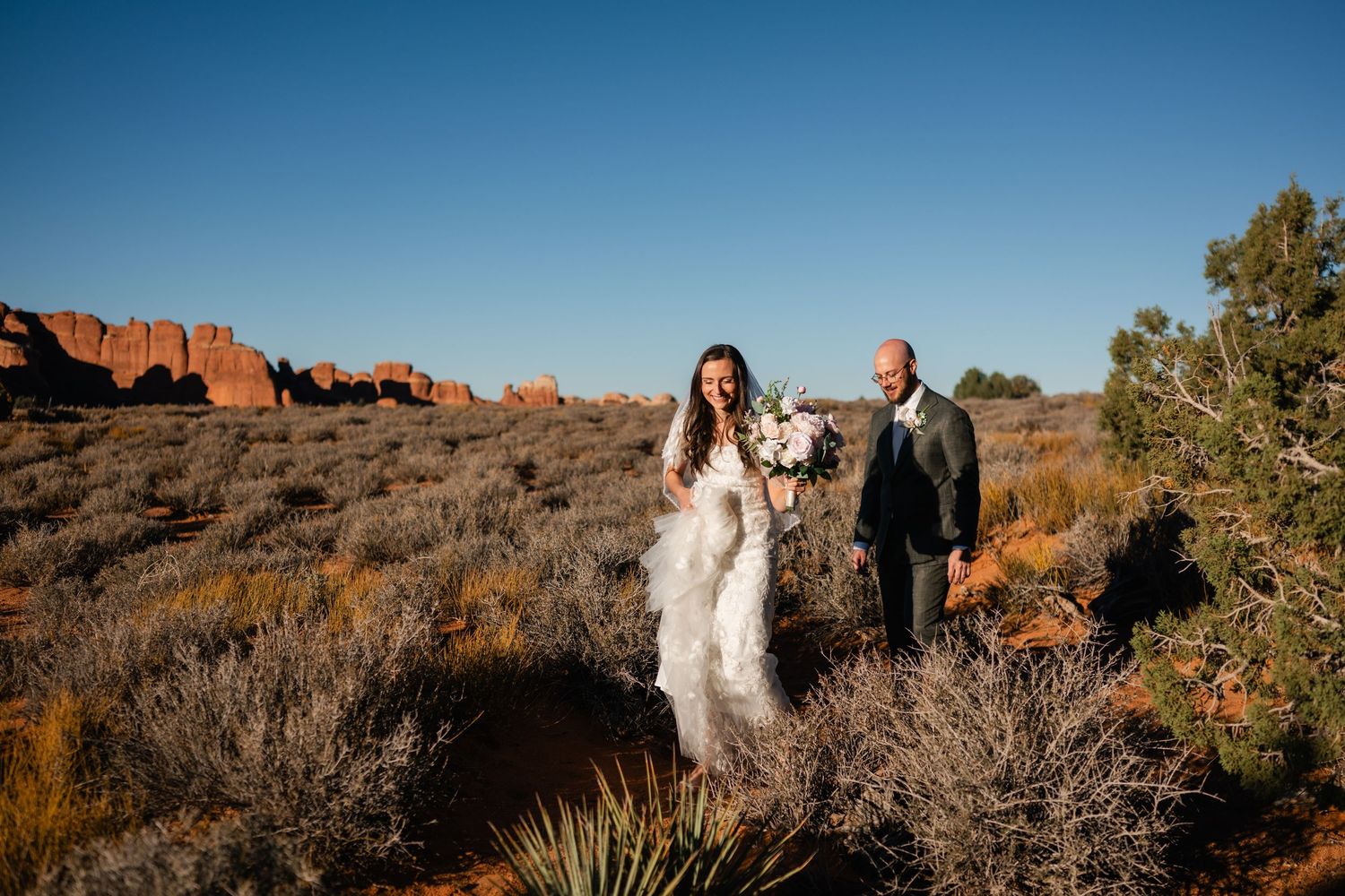 Wedding couple poses against stunning red rock formations in Arches National Park during golden hour.