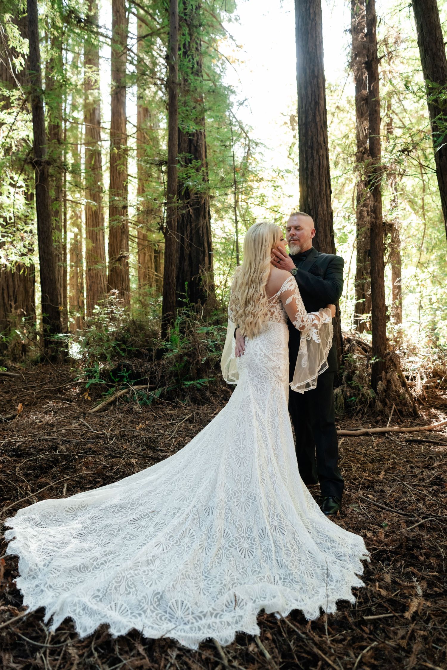 A flowing white lace wedding dress with long train spreads beautifully across forest floor.