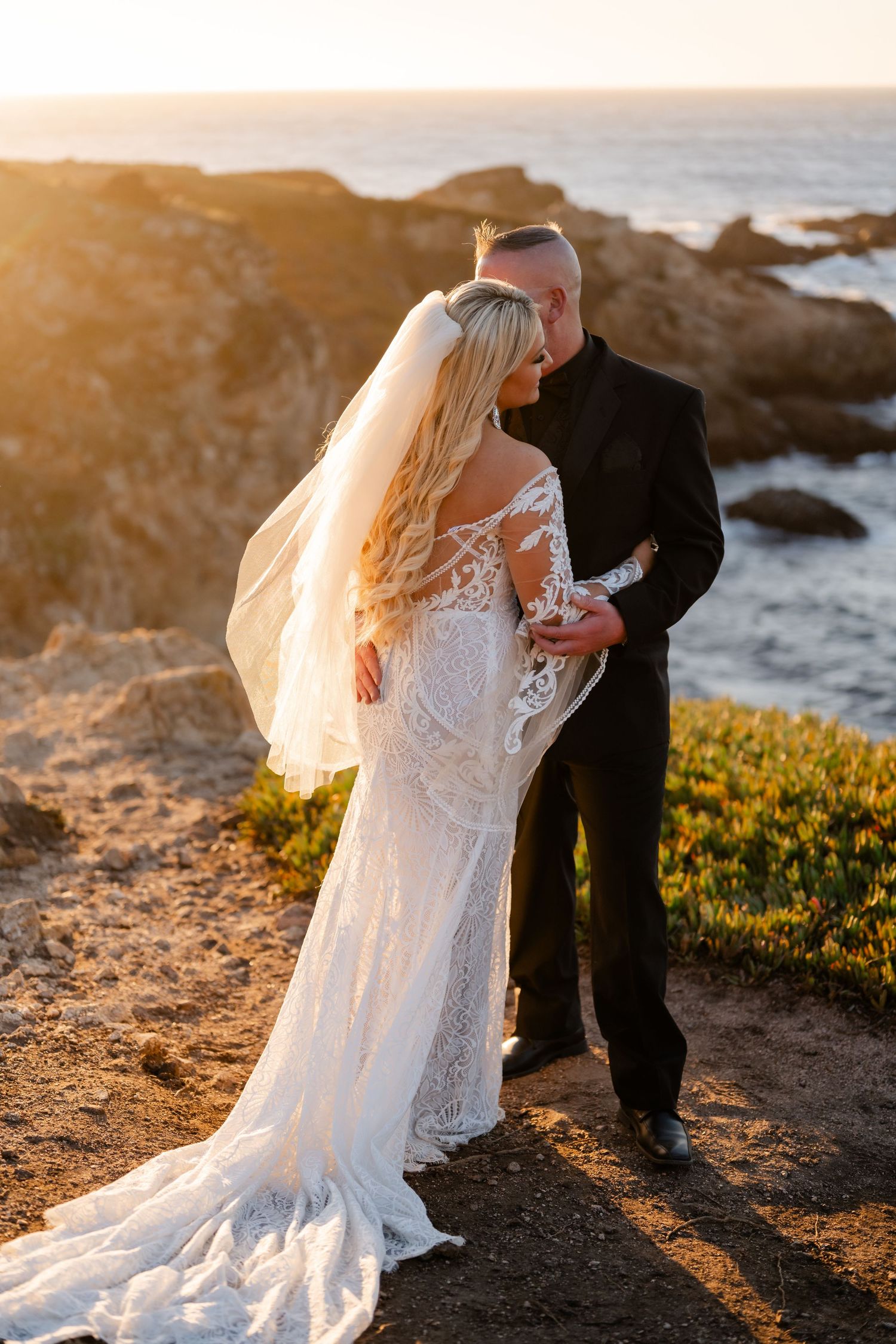 Couple in wedding attire sharing intimate moment on coastal cliffs at sunset.