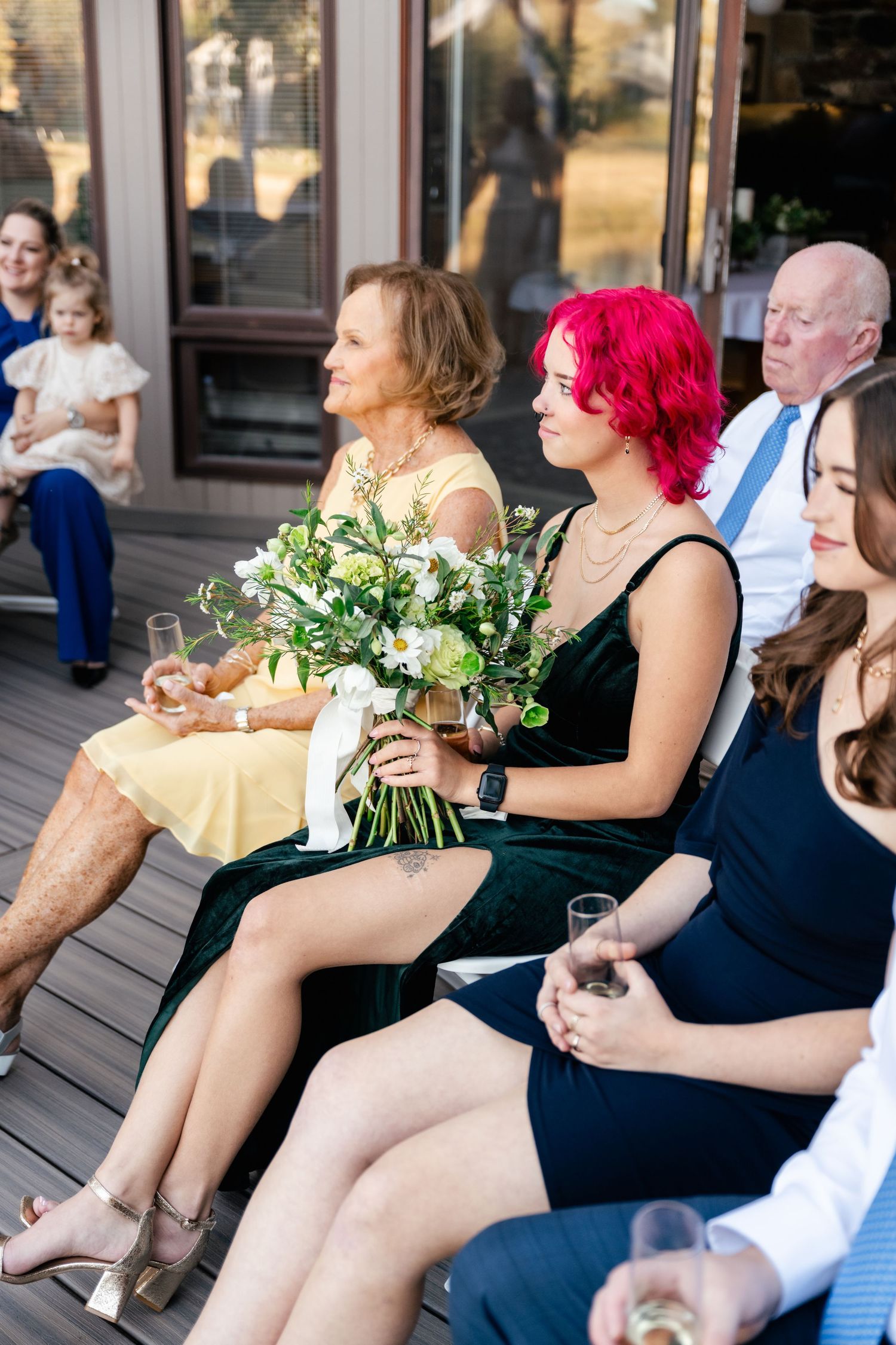 Guests seated on a deck holding drinks and flowers during an outdoor event.