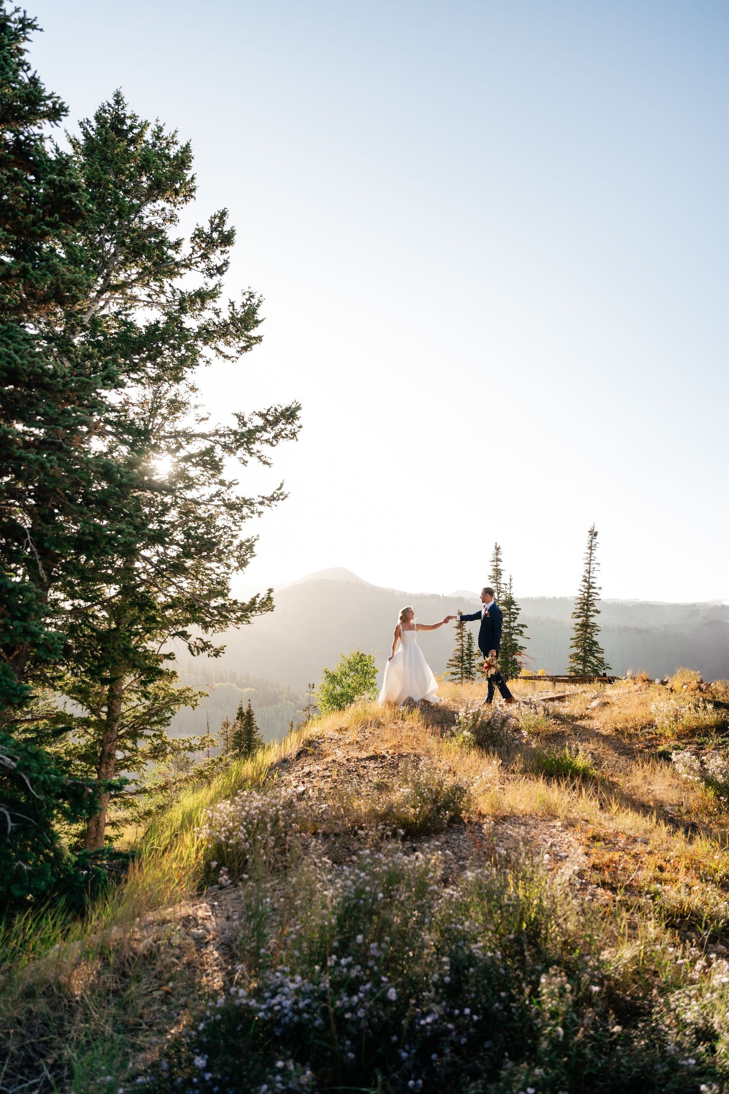 Wedding couple on a scenic hilltop at sunset with mountains and evergreen trees in the background. Park City Wedding Photographer.