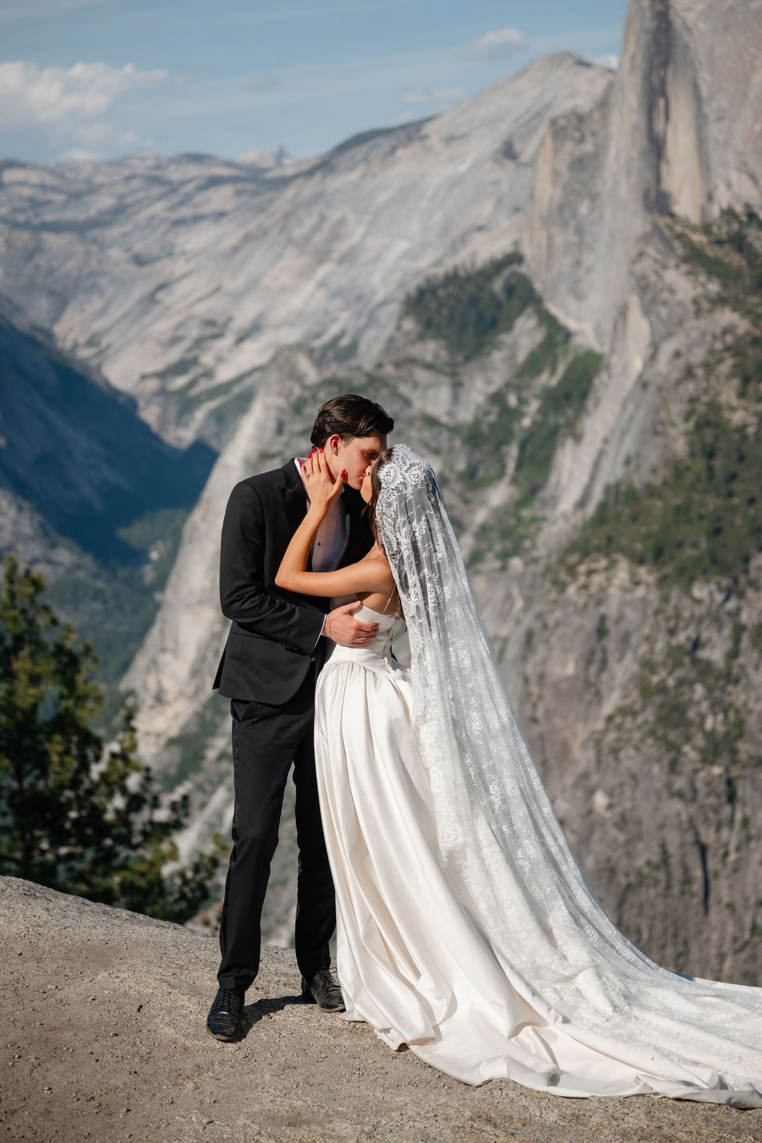 Bride and groom share a kiss on rocky overlook with stunning Yosemite valley and Half Dome behind them.