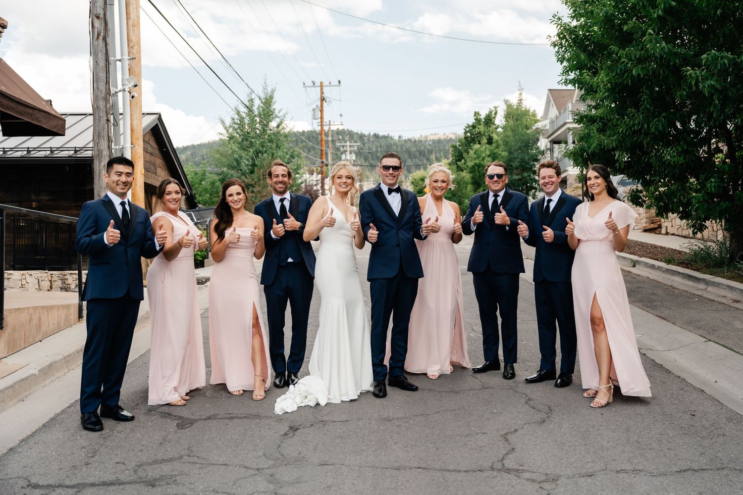 Wedding party in navy suits and blush gowns giving thumbs up on a street with scenic mountain backdrop.