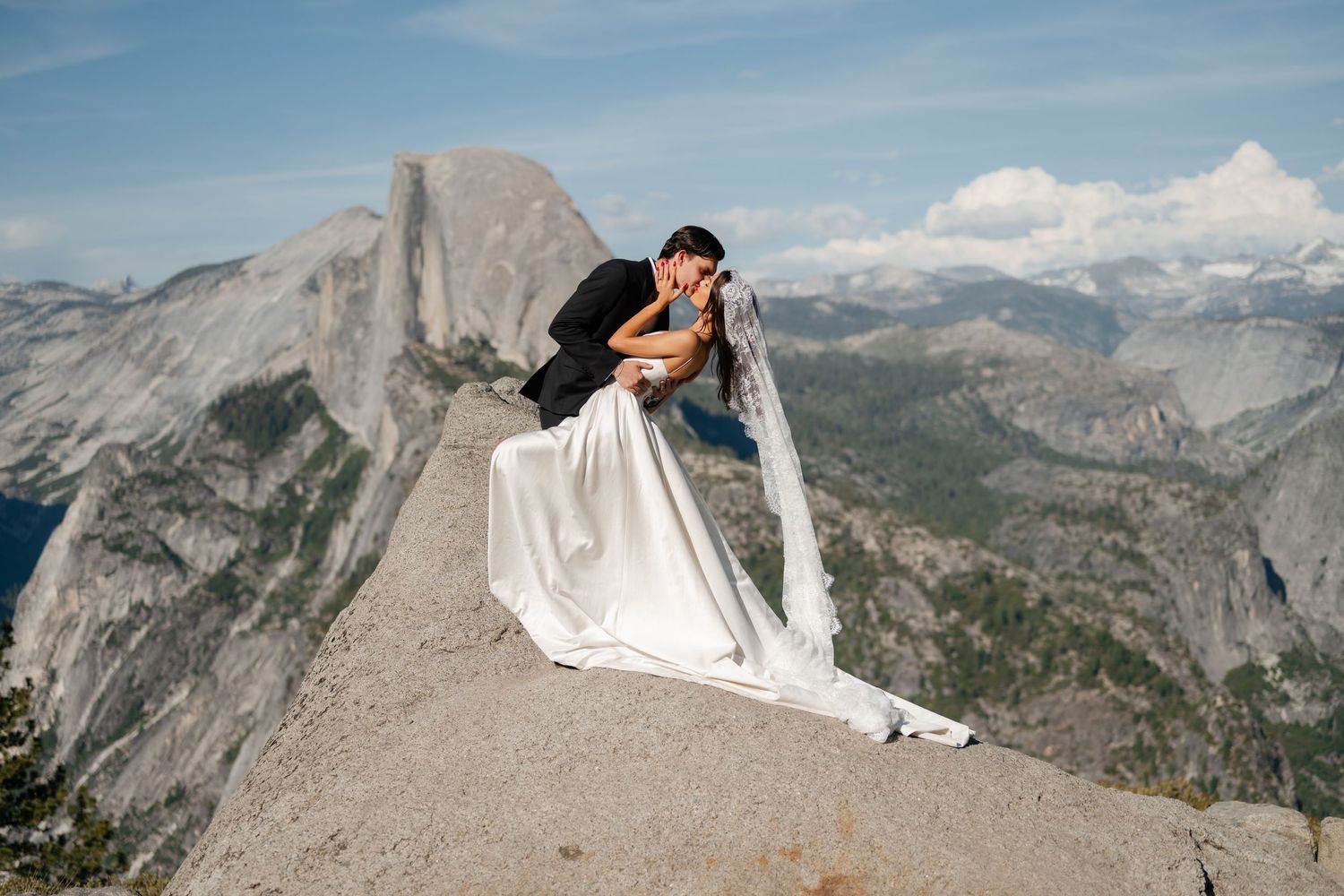 Couple sharing a romantic dip kiss on a rocky summit at Yosemite with Half Dome and valley views behind them.