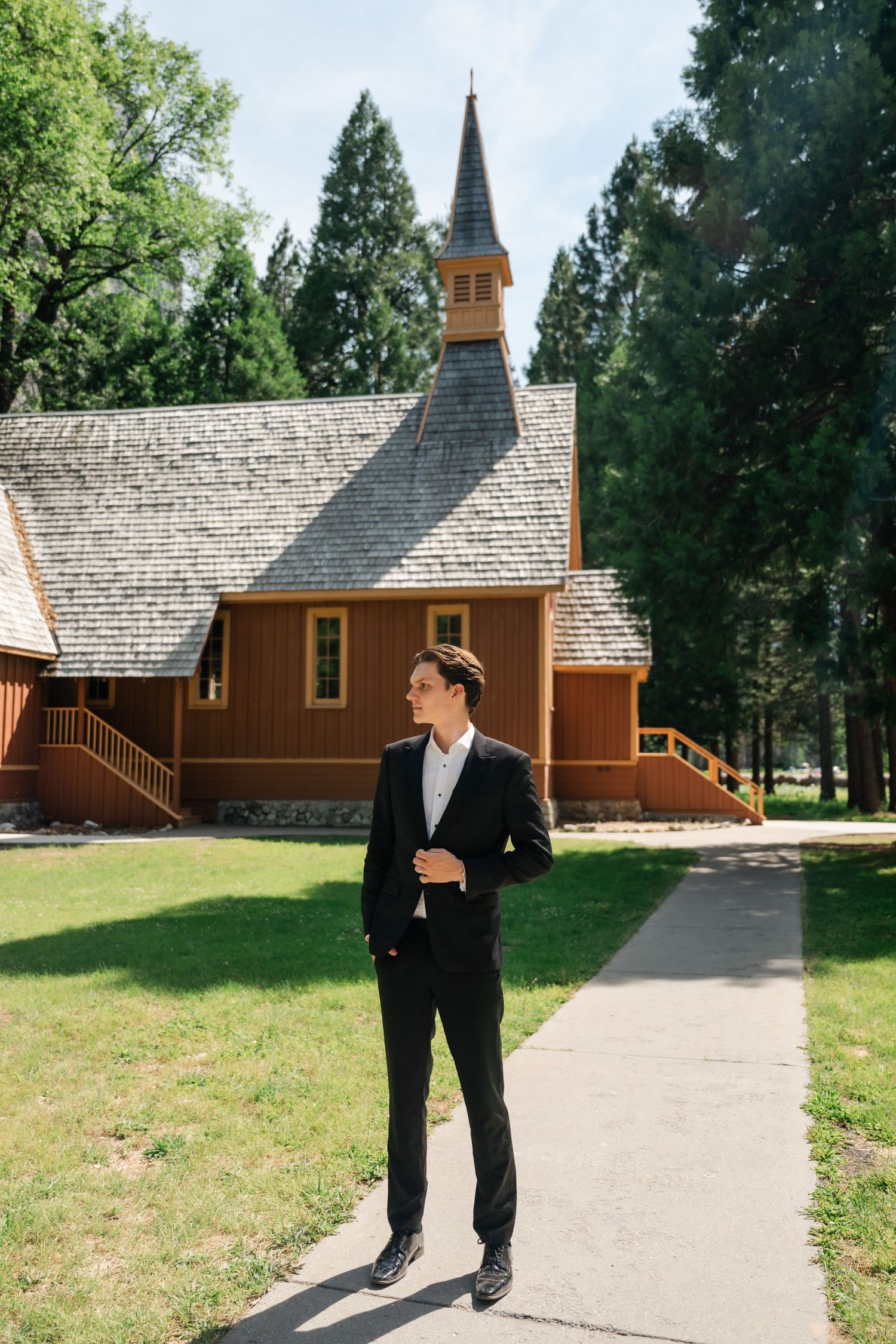 Groom in black suit stands on pathway before rustic wooden chapel with steeple surrounded by tall trees.