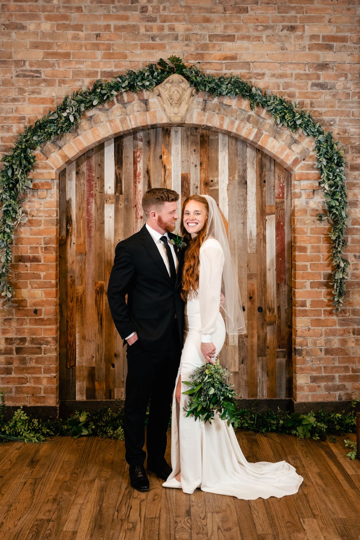 A romantic wedding photo with a greenery arch and wooden door backdrop on a rustic brick wall.