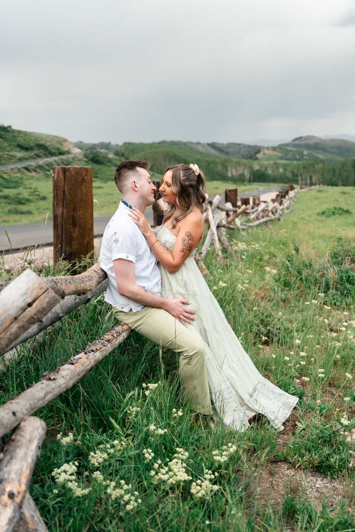 A couple shares a romantic moment sitting on rustic wooden fence posts in a green mountain meadow. Park City engagement photographer.