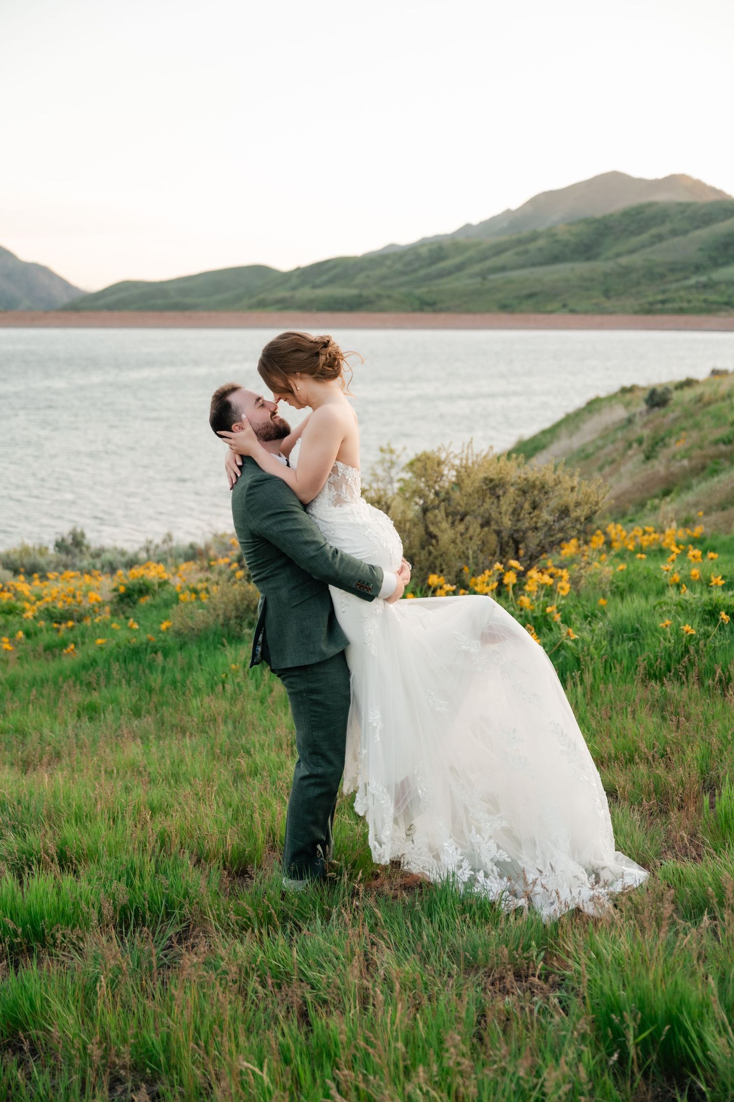 Wedding couple embraces on hillside overlooking lake with wildflowers and mountains in background.